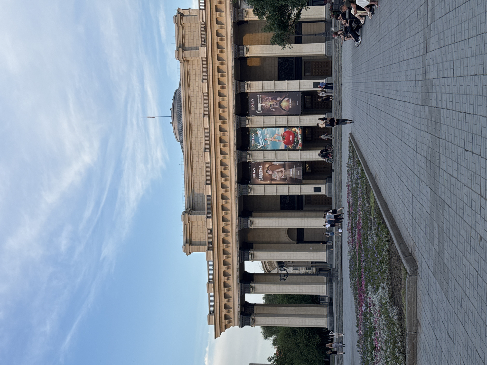 The image shows the Novosibirsk Opera and Ballet Theatre in Novosibirsk, Russia. The theatre is a large, imposing building with a neoclassical facade featuring numerous columns and ornate detailing. Several large banners are hanging from the facade, advertising upcoming performances. In front of the theatre, there is a wide paved plaza with a flowerbed in the foreground. Several people are visible in the plaza, some walking, some sitting on benches, and a few near the entrance of the theatre. The sky above is blue with some wispy clouds. The scene appears to be set during the late afternoon or early evening, with soft lighting.