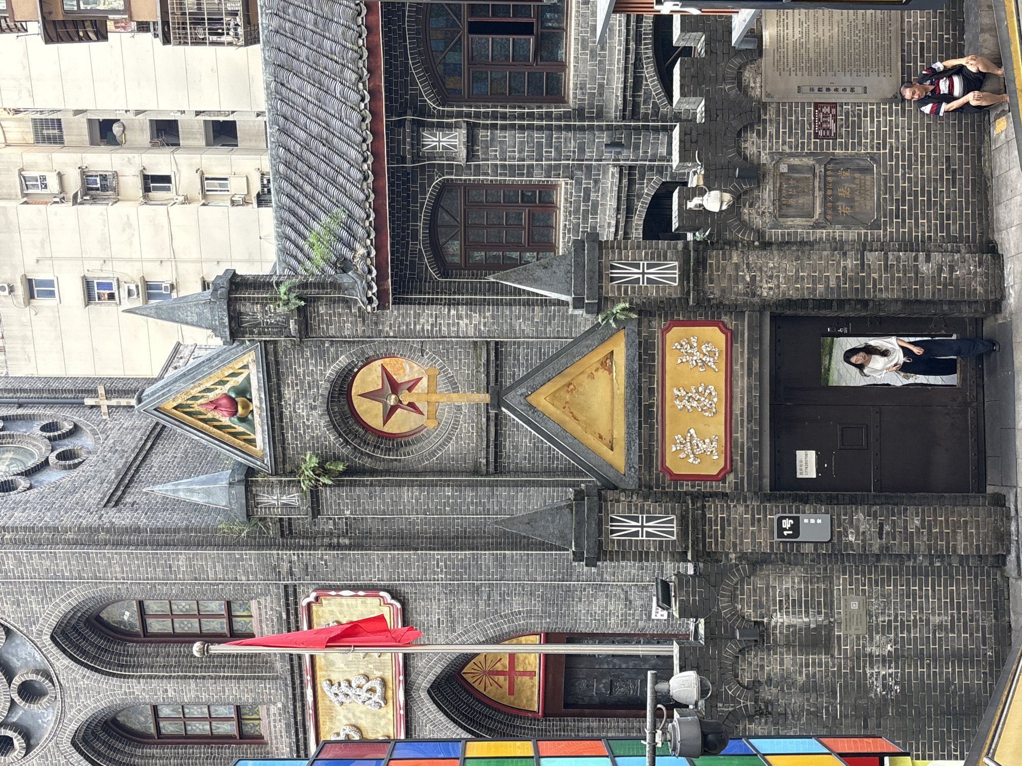 The image shows the exterior of a historic brick building, likely a church or religious institution, in Yuzhong District, China. The architecture features arched windows with stained glass, a prominent red star with a halo above a crucifix, and a tiered facade with crenellated battlements. A red flag is displayed on a flagpole to the left of the entrance.
Two individuals are visible: a woman dressed in a white top and dark pants stands in the open doorway, and a man in a striped vest and shorts sits on a step to the right. The building is surrounded by other urban structures, including modern high-rise buildings in the background. The scene appears to be a street-level view, with a paved walkway in the foreground.