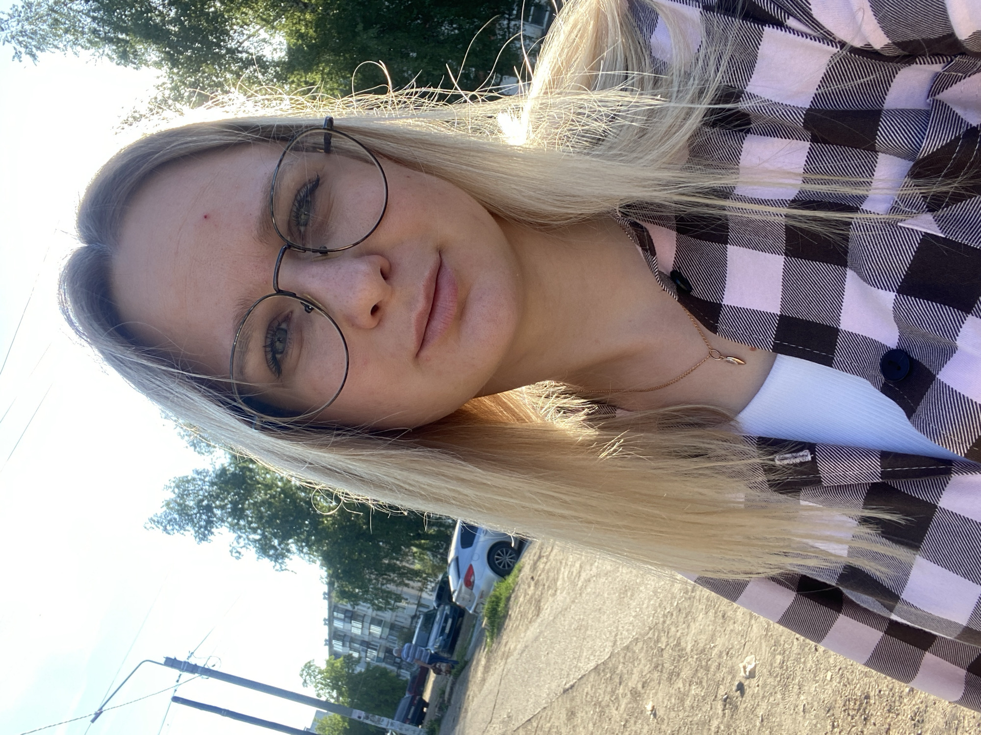 A woman with blonde hair and glasses is standing outside on a sunny day. She is wearing a white top and a pink and black checkered shirt. There are trees and buildings in the background, as well as cars parked on the street. The ground is paved.