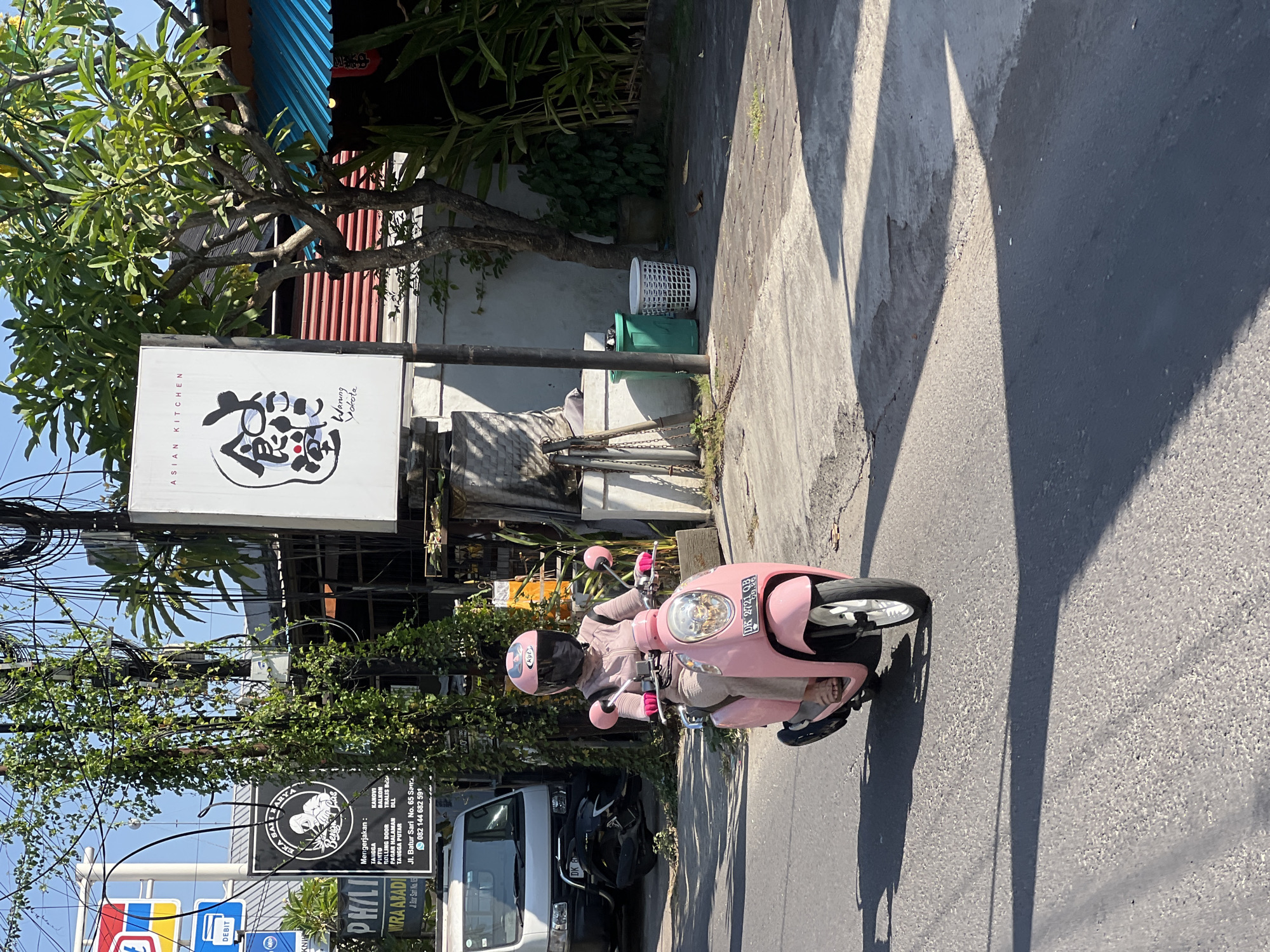 A woman wearing a pink helmet, face mask, and jacket, along with beige pants and flip-flops, rides a pink scooter on an asphalt road in Denpasar, Indonesia. She is heading towards the right side of the frame. The scooter's license plate reads "DK 2721 QB". On the left side of the road, a white van is parked, and behind it, a black scooter is partially visible. Adjacent to the van is a white sign with text in Indonesian and a logo of a person holding a tool. Further left, a blue and red sign with the word "DEBIT" is affixed to a pole. Above the van, a dense network of electrical wires is strung between poles, with vines growing around one of the poles. To the right of the road, a white sign with "ASIAN KITCHEN" and Japanese calligraphy is displayed. Next to this sign, a tree with green foliage grows, and a building with a blue-tiled roof is visible. In the foreground, the road shows patches of wear and tear, and long shadows are cast by the objects on the left. Two trash bins, one green and one white, are situated on the right side of the road. The weather appears sunny with clear skies.