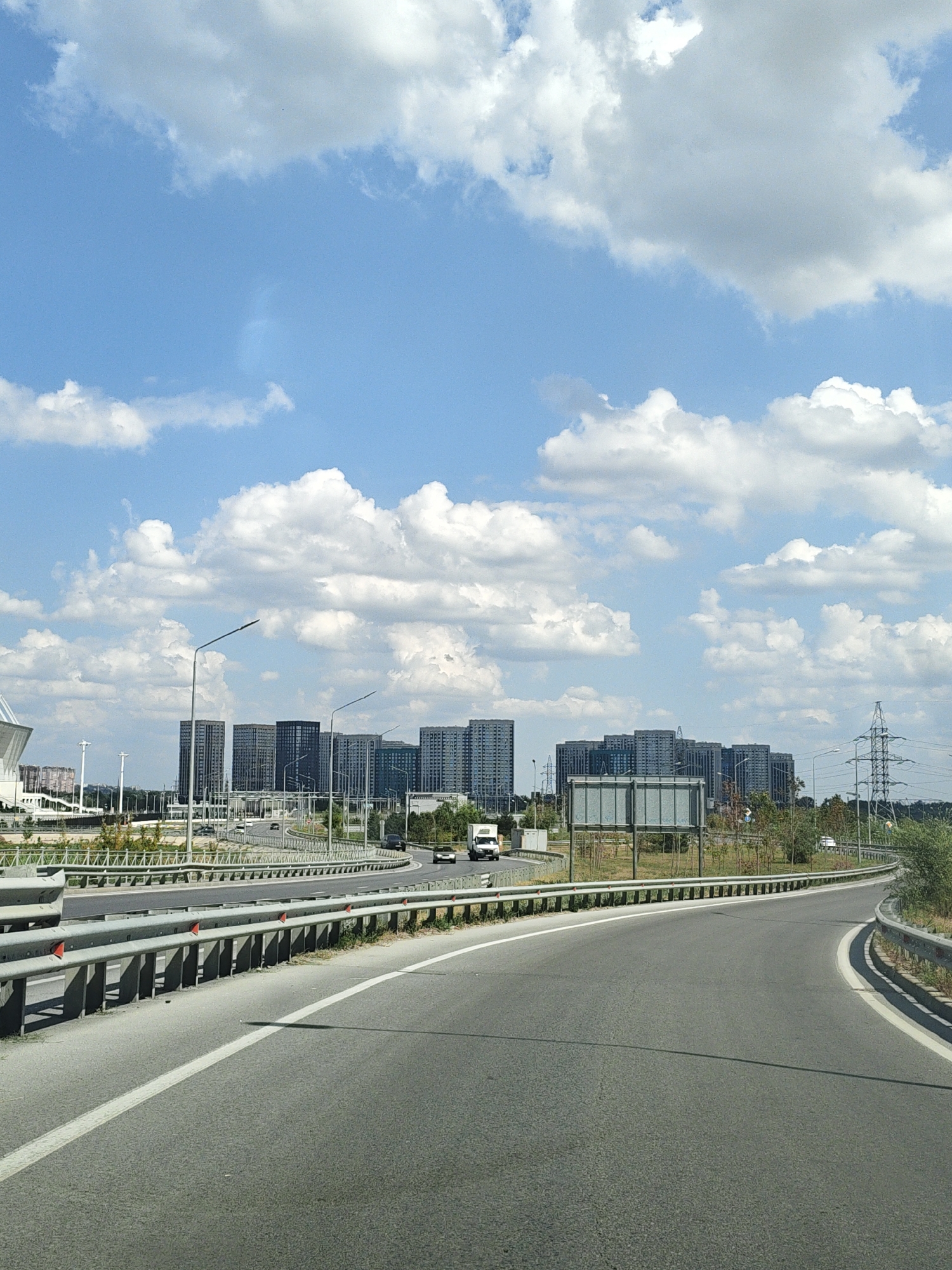 The image depicts a view from a vehicle traveling on a highway. The highway curves to the right, with guardrails separating the lanes. In the background, a cluster of modern, multi-story buildings forms a cityscape. Several tall lampposts line the highway. Power lines and transmission towers are visible on the right side of the road. The sky is blue with fluffy white clouds. The scene appears to be on the outskirts of a city, possibly Rostov-on-Don, Russia, given the architectural style of the buildings and the infrastructure. There are no people visible in the image. Vehicles, including a white truck, are present on the highway. The environment is an urban or suburban highway setting.