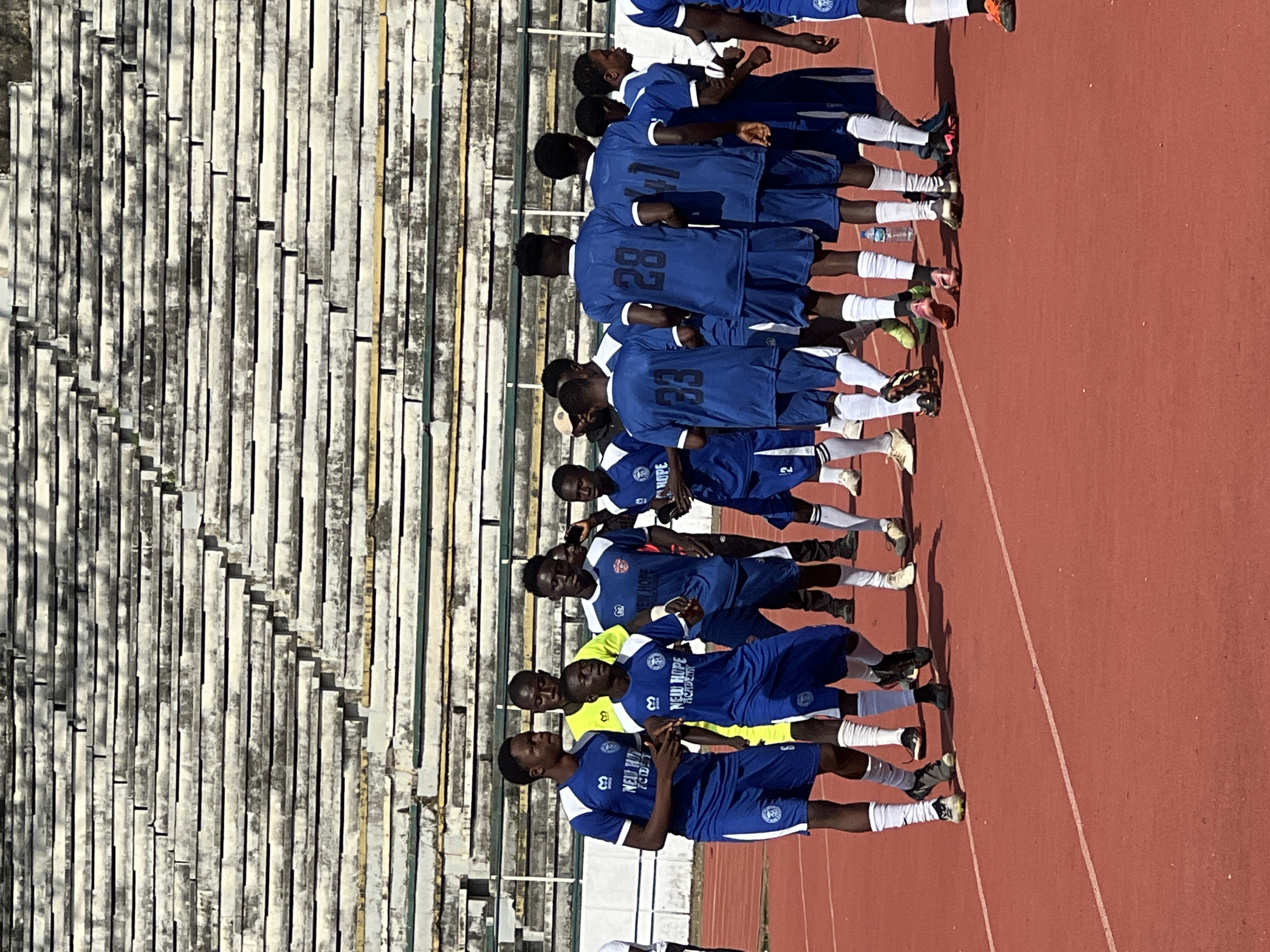 In the city of New Stadium, Nigeria, a group of young male soccer players, dressed in blue and white uniforms with "New Hope Academy" printed on them, are gathered on a red running track in front of tiered concrete stadium seating. The players appear to be in a huddle or discussion, with some looking towards the camera and others interacting with each other. Their expressions are neutral. The background consists of the weathered, empty stadium seats, suggesting the team is either preparing for a match or has just concluded one. The lighting indicates it is daytime, with shadows cast on the track.