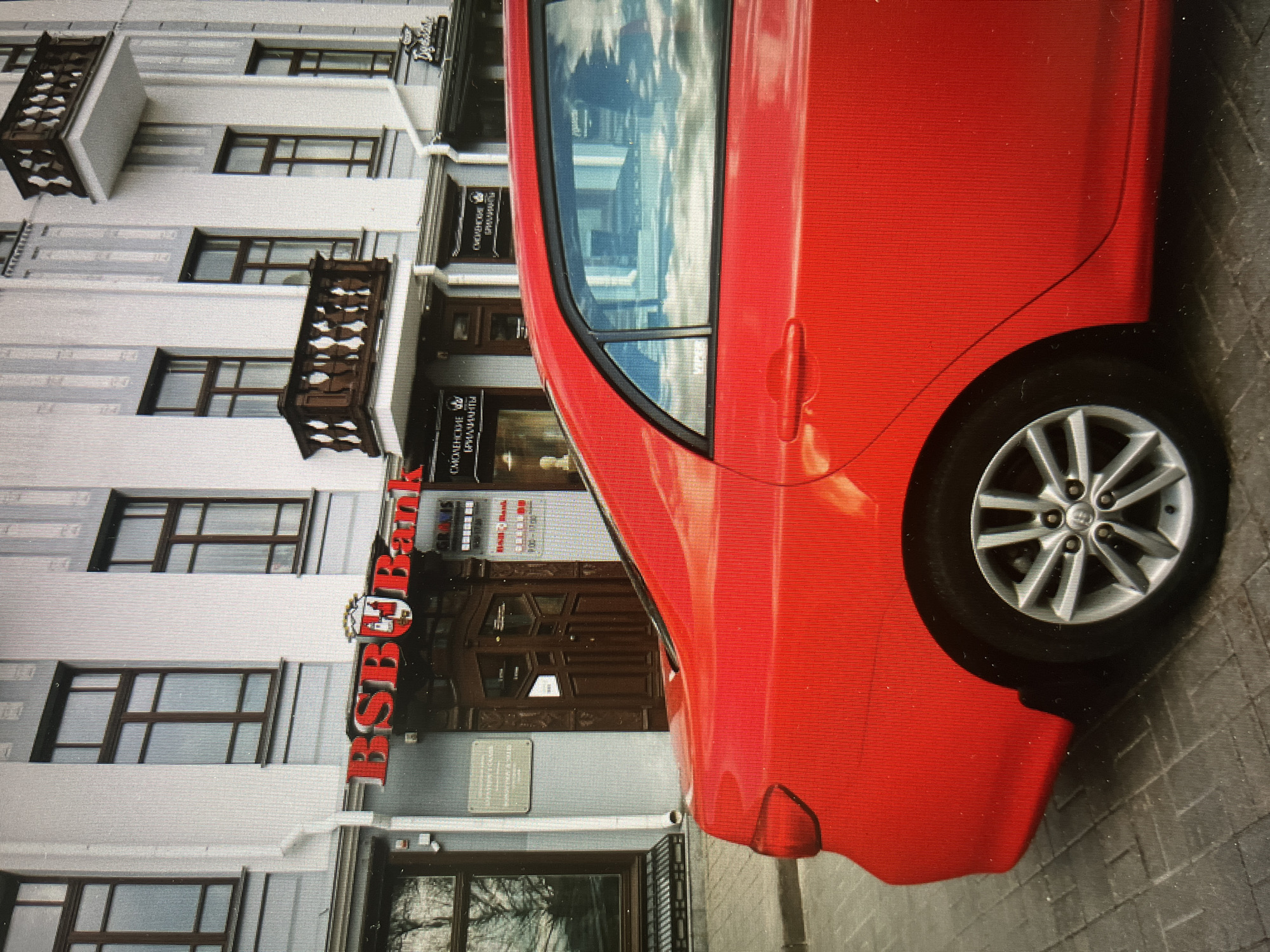 The image shows a red car parked in front of a building with "BSB Bank" signage in Minsk, Belarus. The building has a light grey facade with multiple windows and balconies. The car occupies the right side of the frame, with its rear portion visible. The "BSB Bank" sign is prominently displayed above the entrance of the building. The background reflects the city's urban environment. The image is taken at a slight angle, and the overall lighting appears to be daylight.