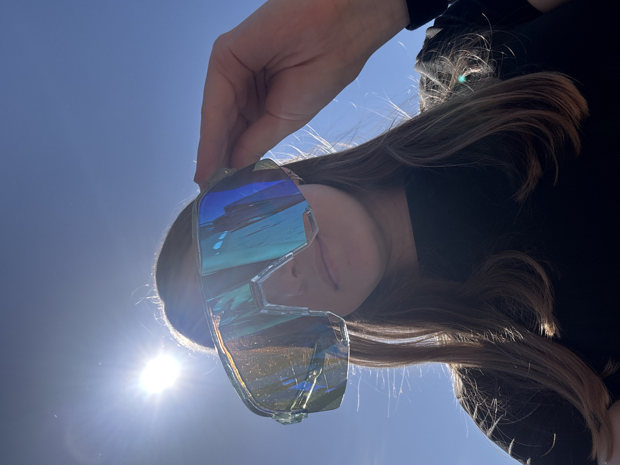A person is holding a pair of iridescent sunglasses up in front of their face. The sky is clear and blue, and the sun is visible in the lower left of the frame. The person has long brown hair and is wearing a black shirt. The sunglasses reflect the blue sky and sun. The location is not specified in the image and I cannot guess this information.