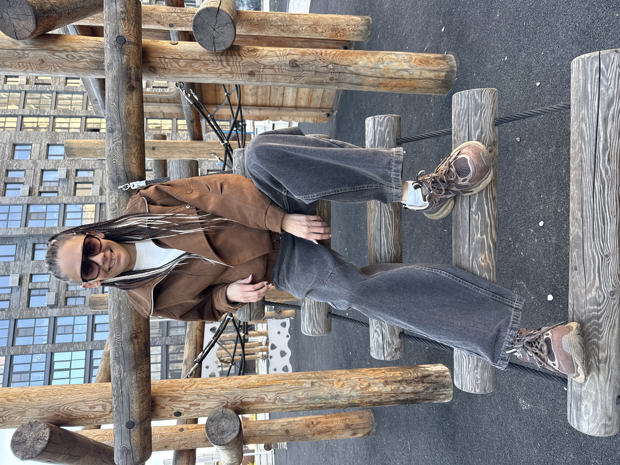 A woman is sitting on a playground structure in Rusanovo, Russia. She has braided hair, a brown jacket, and dark sunglasses. The playground has wooden elements and ropes. A tall building is in the background.