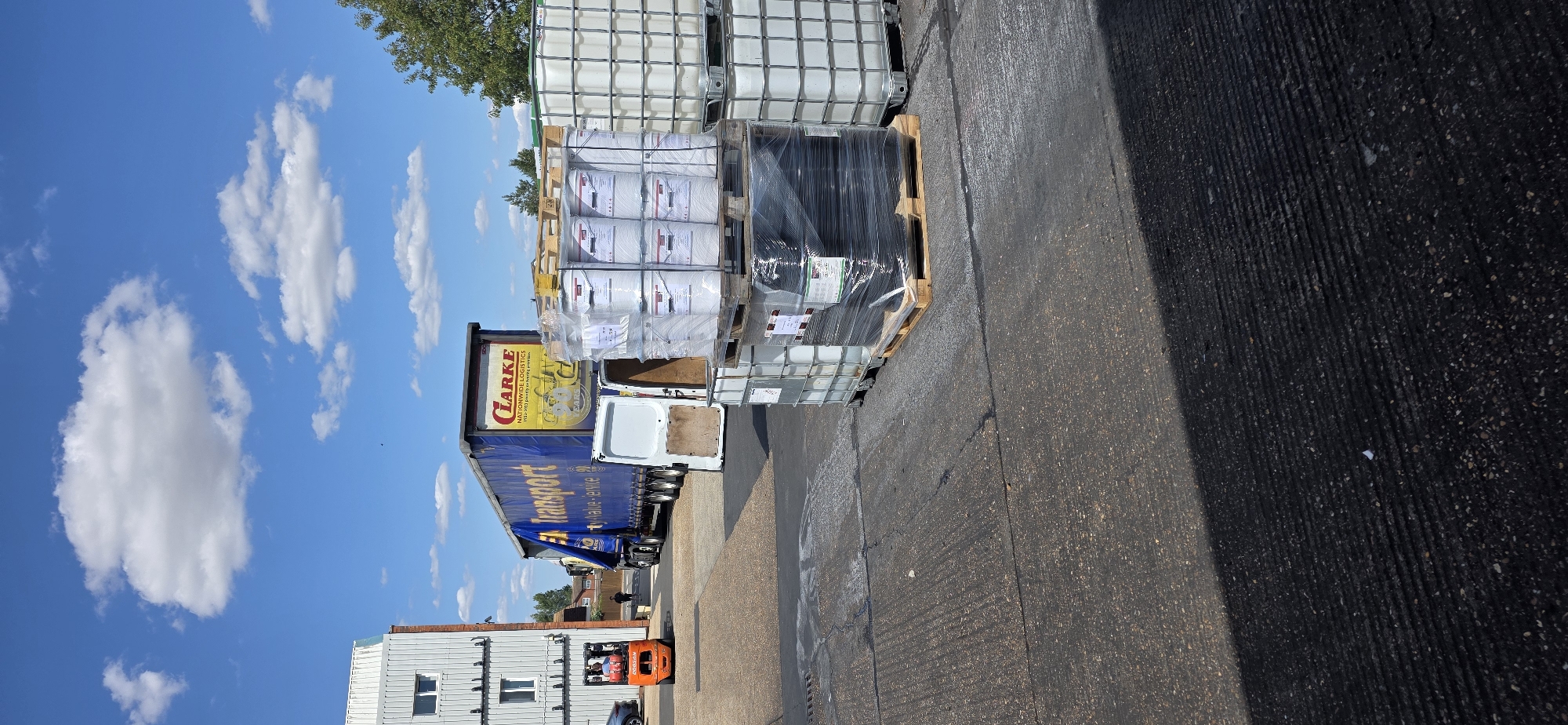 A wide shot captures a loading dock area under a bright, cloudy sky. A large blue and yellow articulated lorry, emblazoned with "CLARKE TRANSPORT NATIONWIDE LOGISTICS," is parked, its trailer open, revealing a white van with its rear doors ajar. In the foreground, pallets of white and black plastic containers are stacked, some wrapped in plastic. To the left, an orange forklift is in operation, with a person visible on it. A multi-story building with white siding and windows is to the far left. The ground is paved with asphalt. This scene appears to be in Arun, United Kingdom, suggested by the branding on the lorry.