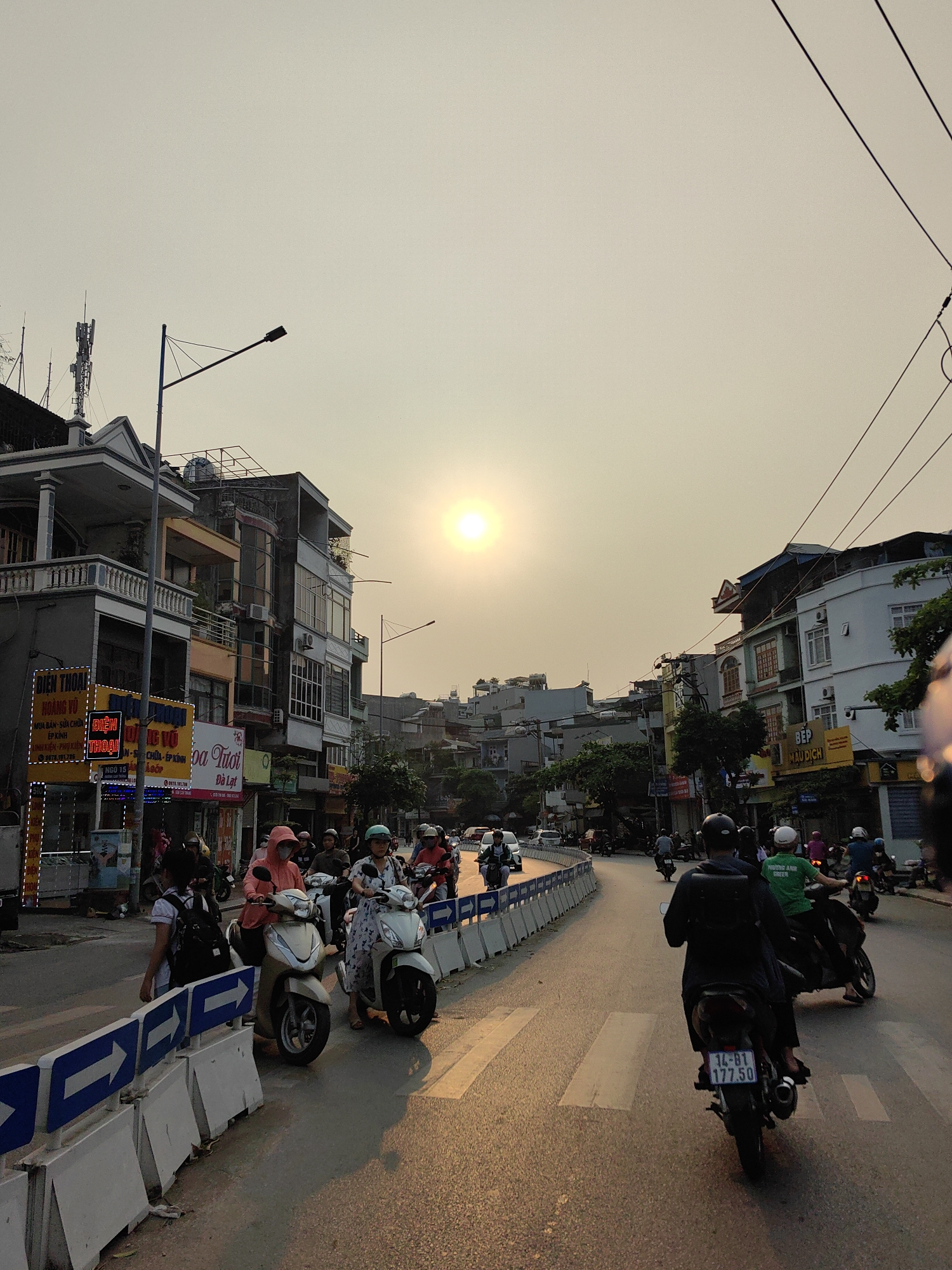 The image captures a street scene in Ha Lam Ward, Vietnam. The street is filled with various motorbikes, some parked and some in motion. People are visible on the motorbikes and walking along the street. Buildings line the street, some with visible signage in Vietnamese. The sun is prominent in the sky, creating a warm, golden light across the scene. Concrete barriers are placed along the side of the road. Electrical wires are visible overhead. A street sign on one of the motorbikes reads "14-B1 177.50".