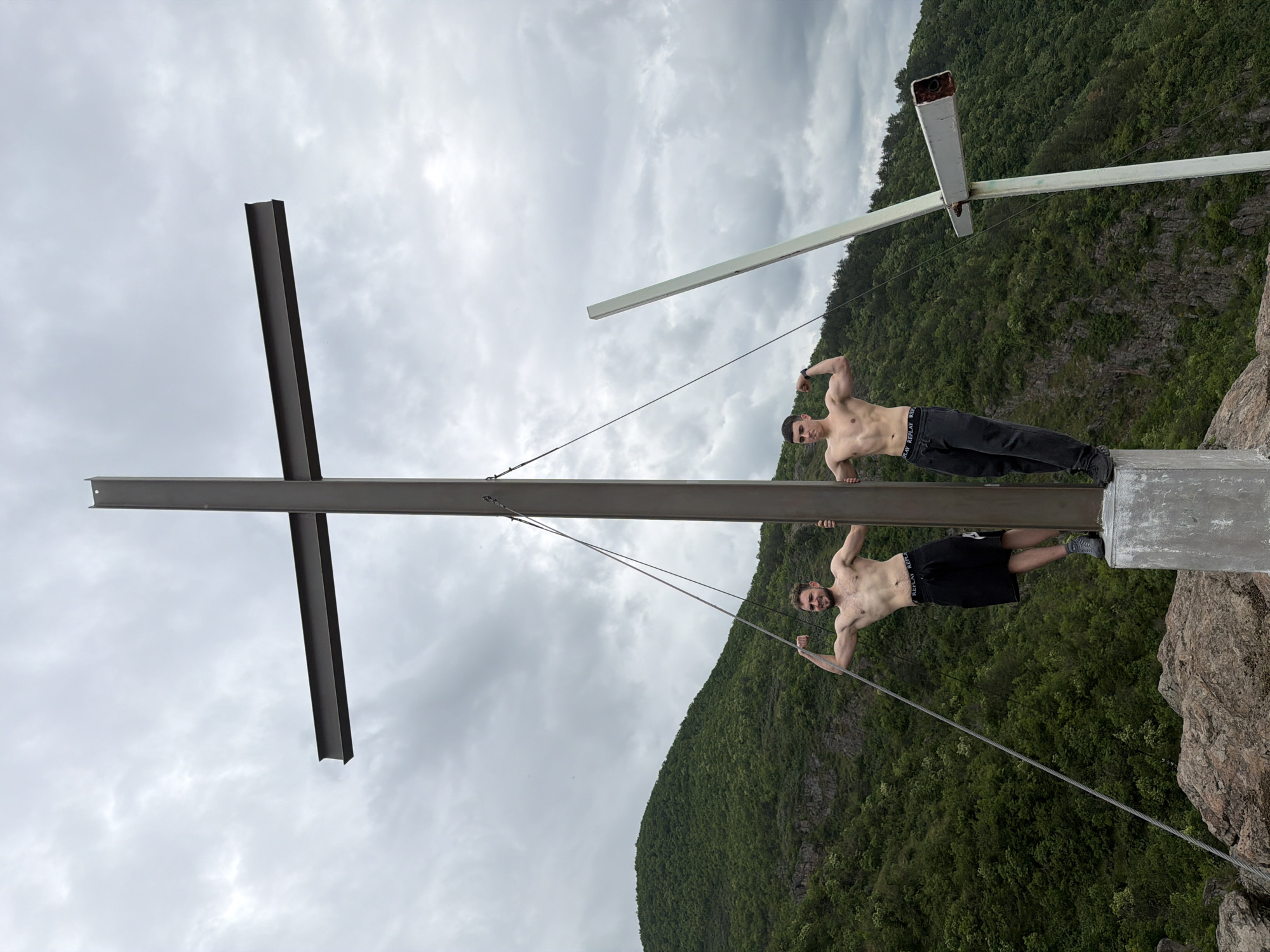 The image shows two men standing on the summit cross of mount Ustina in Bulgaria, which features two conjoined cross structures. Both men are shirtless and appear to be holding onto the cross structure and flexing their arms. One man is wearing black shorts while the other is wearing black pants. The weather appears overcast and the mountain is covered in green foliage.