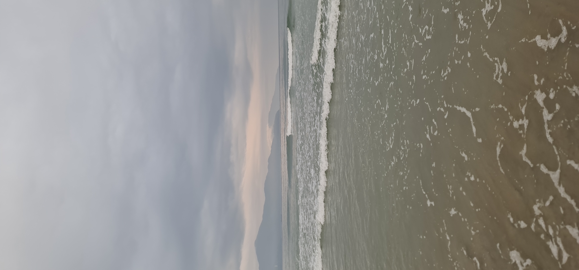 This media file captures a serene coastal landscape in Đà Nẵng, Vietnam, likely during an overcast day, possibly early morning or late afternoon, given the diffused light. The image is composed horizontally, with a vast cloudy sky occupying the upper portion, a distant mountain range forming the horizon, and the ocean with breaking waves in the foreground.
The sky is predominantly light grey with varying shades of darker grey clouds, suggesting an overcast or cloudy weather condition. Towards the horizon, where the sky meets the distant landmass, there's a subtle band of lighter, softer hues, possibly indicating a hint of sunrise or sunset colors struggling to break through the cloud cover, casting a delicate, diffused glow.
A range of mountains or hills stretches across the middle distance, appearing dark and hazy against the lighter horizon, their forms softened by atmospheric perspective. These landforms delineate the coastline.
The majority of the image is dominated by the ocean. The water surface displays gentle waves, with white foam and turbulent patterns indicating the movement of the surf. Closer to the viewer, the water has a muted greenish-brownish tone, with numerous patches and streaks of white foam from receding waves. Further out, the ocean appears a more uniform grey-green. The waves are relatively calm, creating a rhythmic pattern of white crests and foamy trails.
No people, specific objects (other than natural elements), or visible text are present in the image. The overall mood conveyed is one of tranquility and quiet contemplation, characteristic of a natural beach scene under a soft, indirect light.