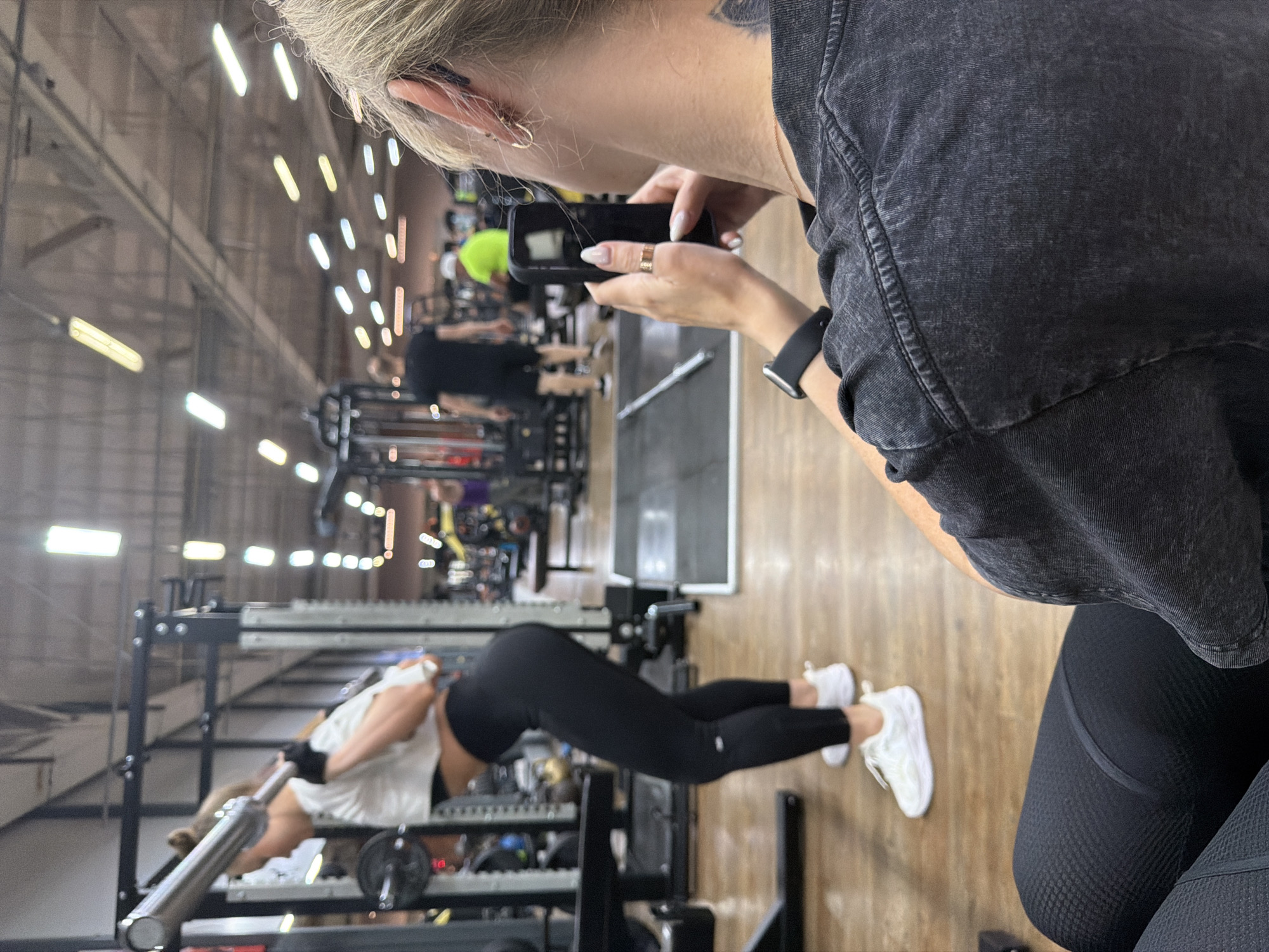 A person is taking a picture with a smartphone of a woman doing a bench press in a gym. The gym has other people and equipment in the background. The image was taken in Chaikovsky, Russia.