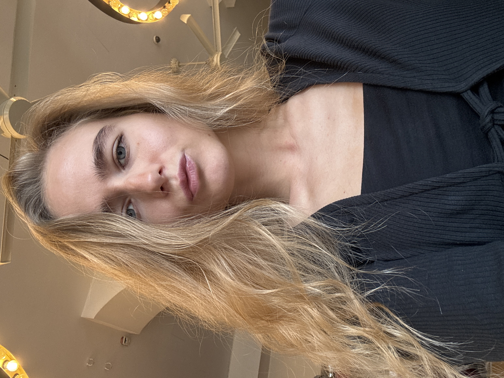 A young, blonde-haired Caucasian woman is centrally featured in this tilted portrait-style photograph. She has fair skin, blue eyes, and lightly tinted lips. Her hair cascades down her right side. She is wearing a black, ribbed top or sweater, partially revealing her neck and shoulder. The background is blurred, suggesting an interior setting. A curved lighting fixture is visible in the upper portion of the image. The photograph has been taken from a low angle, looking up at the woman. The location is specified as Unknown, Unknown.