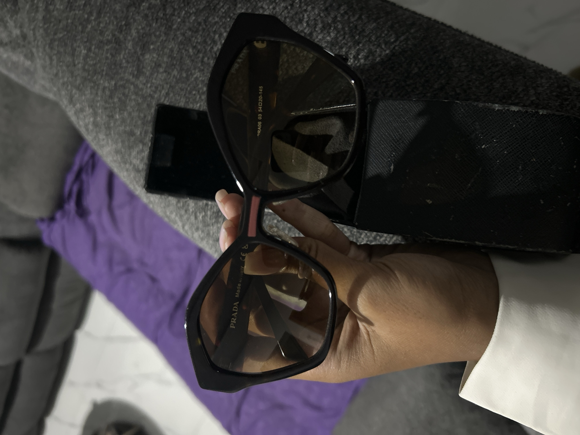 A person's hand holds a pair of black Prada sunglasses with brown lenses. The sunglasses are positioned in front of a dark gray sofa and a purple throw pillow. On the temple of the sunglasses, the text "PRADA Made in Italy CE" is visible. Below that, another line of text reads "PRA06 03 54 20-145." A black sunglasses case is visible behind the sunglasses. The background consists of a blurred sofa and a floor. The image does not provide any information about the city of Sukawati, Indonesia.