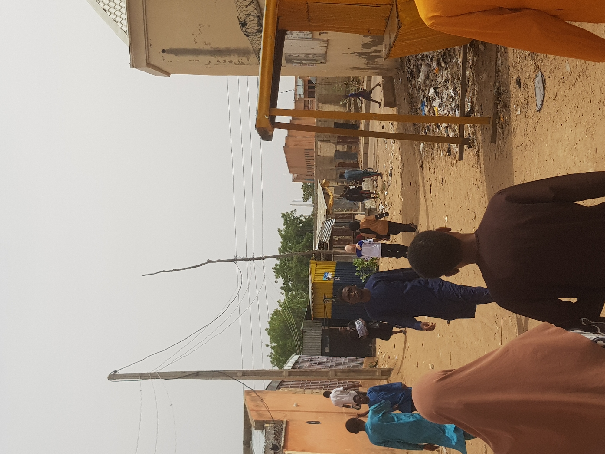 The image depicts a street in Dabawa, Nigeria. Several individuals are walking on the dirt road. Buildings of varying heights and construction materials line the street. A utility pole stands tall with electrical wires extending across the sky. There's debris and litter on the ground. Overhead, a yellow metal structure is visible. A cluster of trees is present in the middle distance.