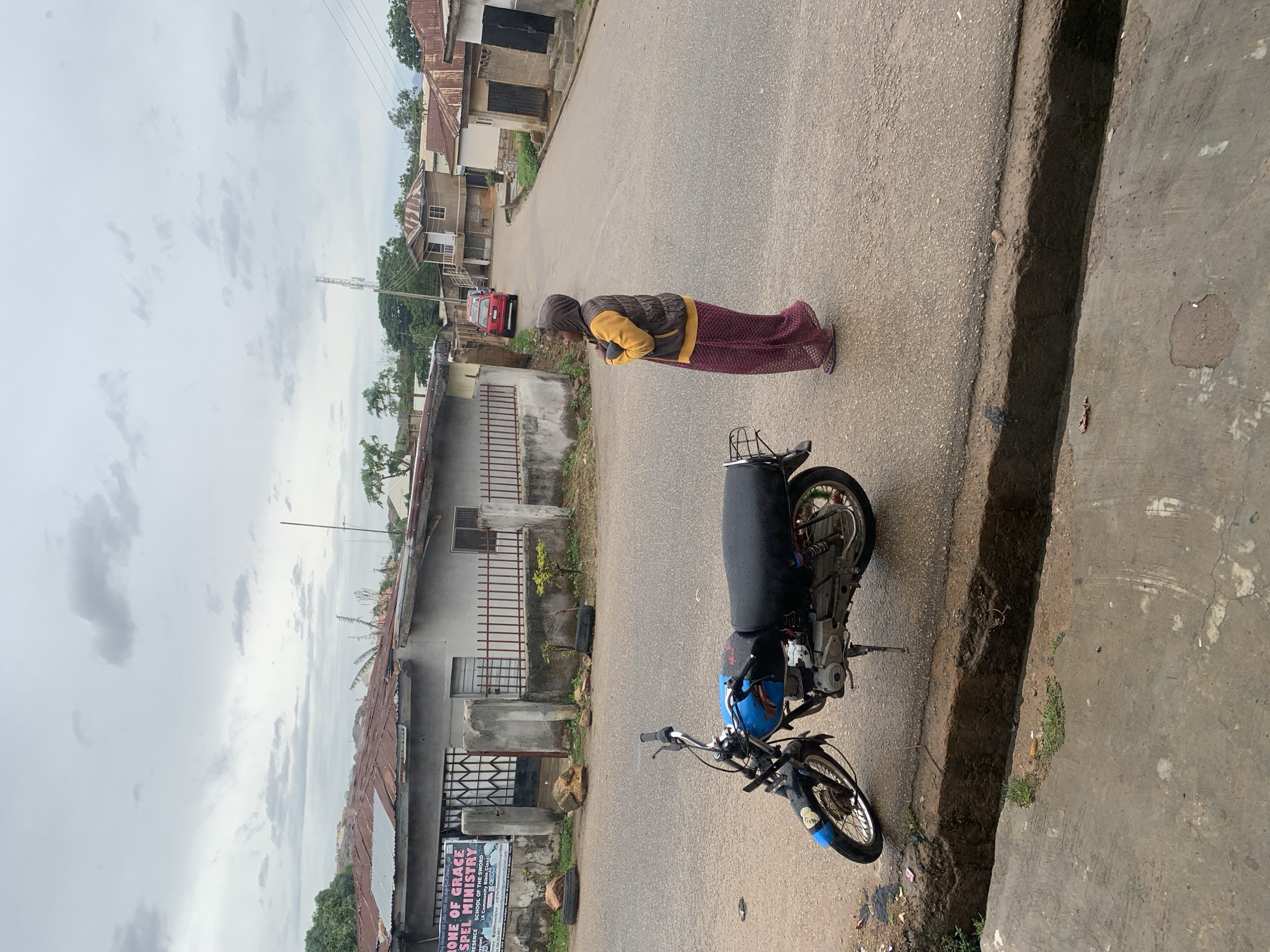 A street scene in Jos, Nigeria, is captured on an overcast day, presenting a view of a residential or semi-urban area. A person, likely a woman, stands on the right side of a paved road, facing slightly away from the camera. She is dressed in maroon patterned trousers or a skirt, a brown vest over a yellow long-sleeved top, and what appears to be a head covering.
In the foreground on the right, a blue and black motorcycle is parked, its seat covered in black material. A concrete gutter runs along the side of the road, separating the pavement from the main thoroughfare. The paved road recedes into the distance, lined on both sides by various single and multi-story buildings, some featuring corrugated iron roofs. A red car is visible further down the street in the midground.
The sky is uniformly grey with clouds, indicating an overcast condition and diffused lighting, suggesting either an early morning, late afternoon, or simply a very cloudy day without direct sunlight. On the left side of the street, a signboard near a building displays the text: "RONE OF GRACE GOSPEL MINISTRY PRESENCE SCHOOL OF THE SWORD A Community Bible Class." The overall impression is a quiet street scene typical of a Nigerian town or city.
