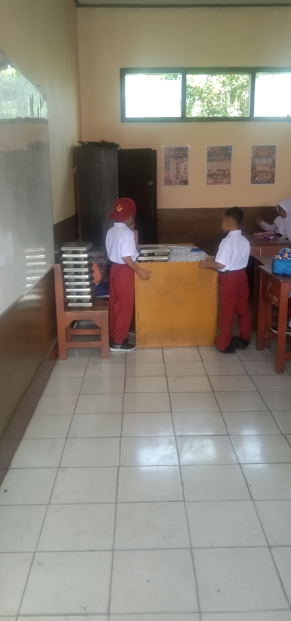 The image shows two young students in uniform, a white shirt and red pants, standing in a classroom. One student, on the left, is wearing a red cap. They appear to be near a teacher's desk. The classroom has a tiled floor, large windows showing greenery outside, and posters on the wall. There is a wooden chair with stacks of trays on it. The overall environment suggests a typical Indonesian elementary school classroom. The image was taken in Rahayu, Indonesia.