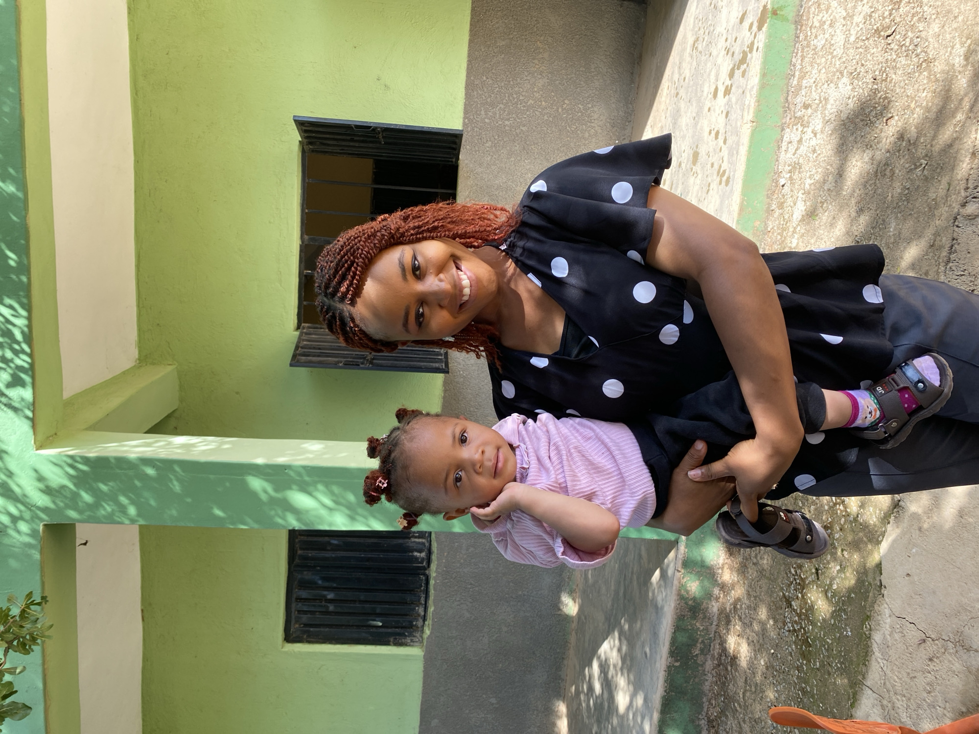 A woman with reddish-brown braided hair holds a young child in her arms. The woman is wearing a black top with white polka dots. The child is wearing a pink striped shirt, black pants, and sandals. They are standing outside a building with a light green painted exterior. The building has black-barred windows. The location is likely Gura Top, Nigeria. The setting is outdoors on a sunny day.