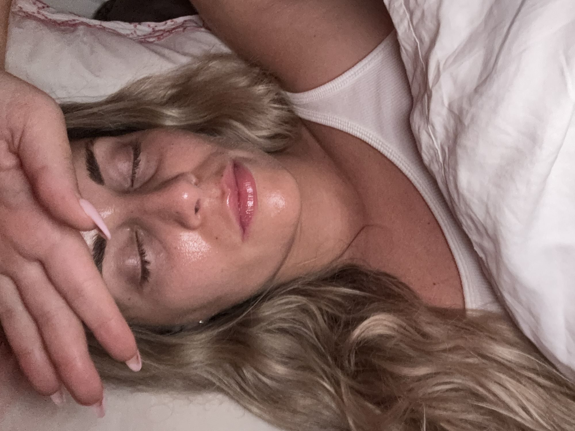 A woman is lying in bed with her eyes closed and her hand resting on her forehead. Her hair is blonde and wavy, and she is wearing a white tank top. The bed is covered with white sheets. The scene appears to be a bedroom. There is no indication that this image was taken in Paris, France.