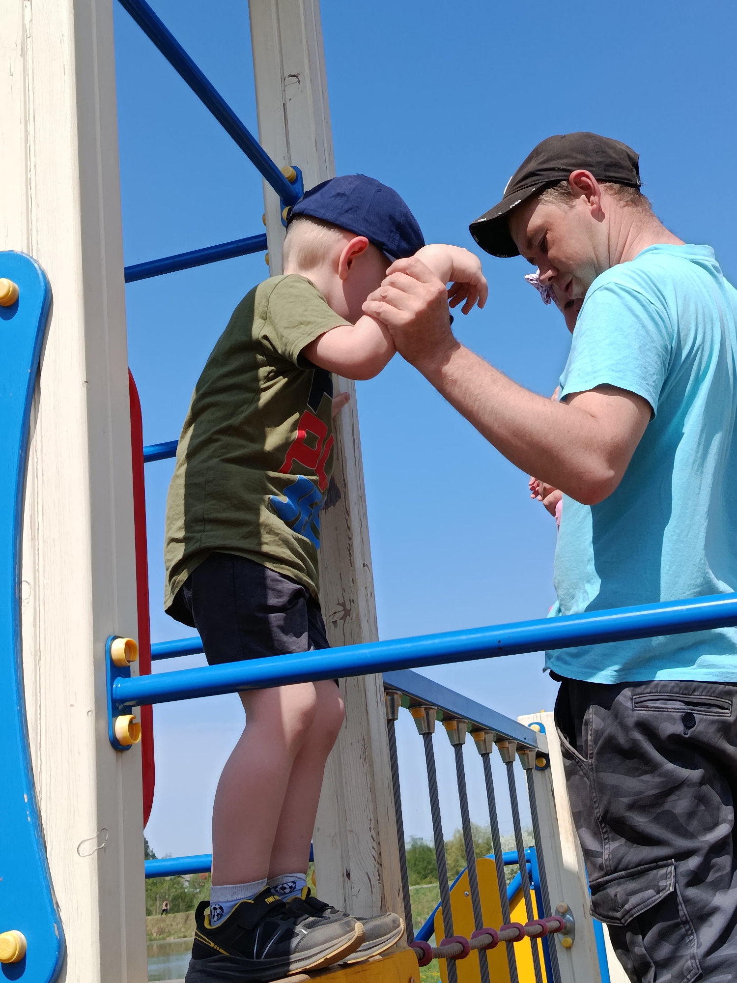 A low-angle shot shows a man and a boy on playground equipment against a clear blue sky. The boy, wearing a navy baseball cap, an olive green shirt with a colorful design, black shorts, and black sneakers with light grey socks, is positioned on a wooden platform, holding onto a wooden beam with blue painted horizontal bars. The man, wearing a black baseball cap, a light blue t-shirt, and camouflage cargo pants, stands to the right of the boy, holding the boy's arm to assist him. The playground structure consists of a wooden frame with blue and yellow accents, blue horizontal bars, and a rope bridge visible below. Trees are visible in the background. The location is Фоки, Russia.