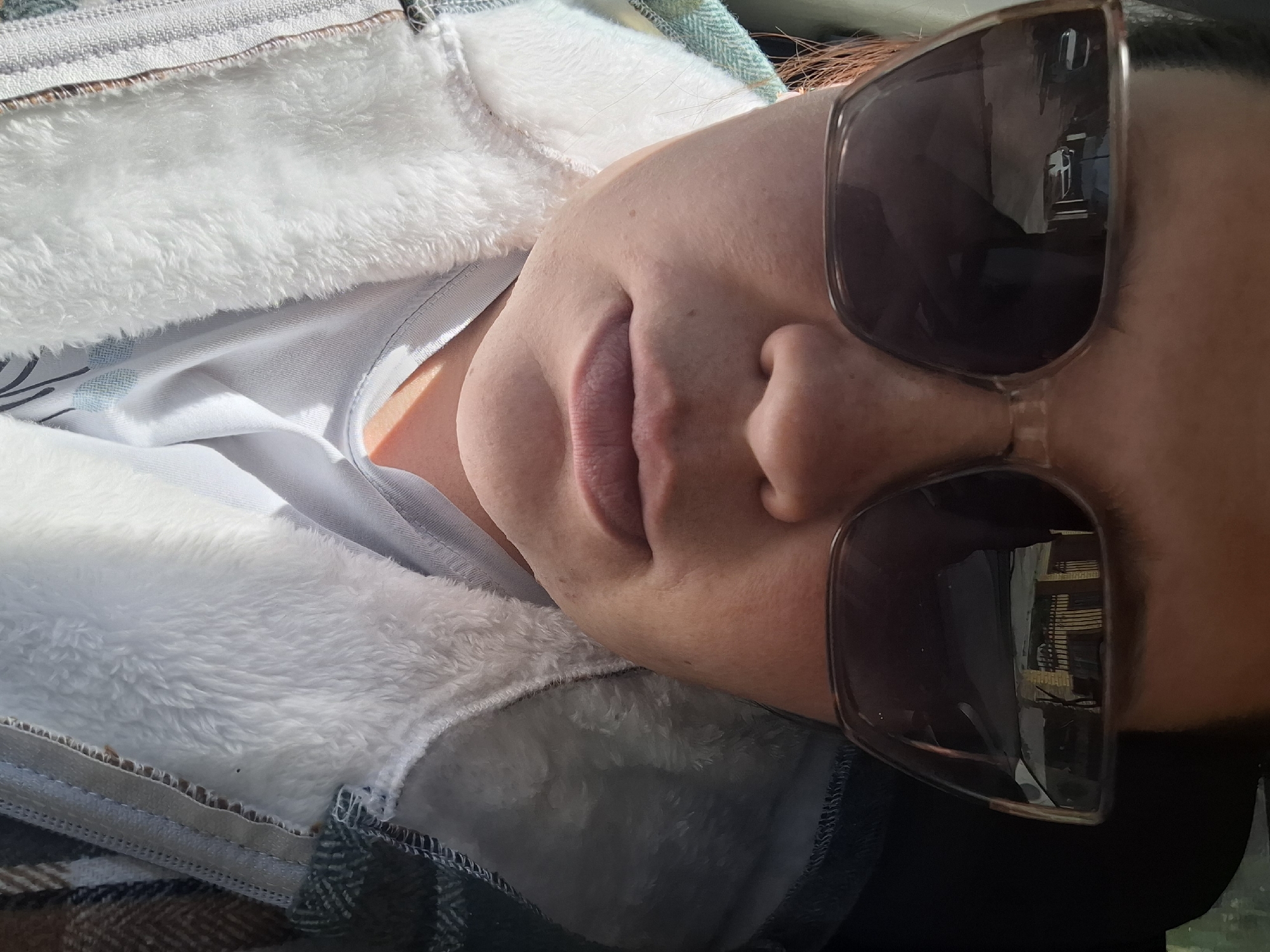 A close-up, somewhat angled portrait shows a person wearing sunglasses. The reflection in the lenses suggests an exterior view of a building, specifically, wooden siding of a home or shed. The person is wearing a white fluffy jacket over a light-colored shirt. A plaid blanket or similar fabric is visible below the jacket. The location is Chaikovsky, Russia based on the reflection.