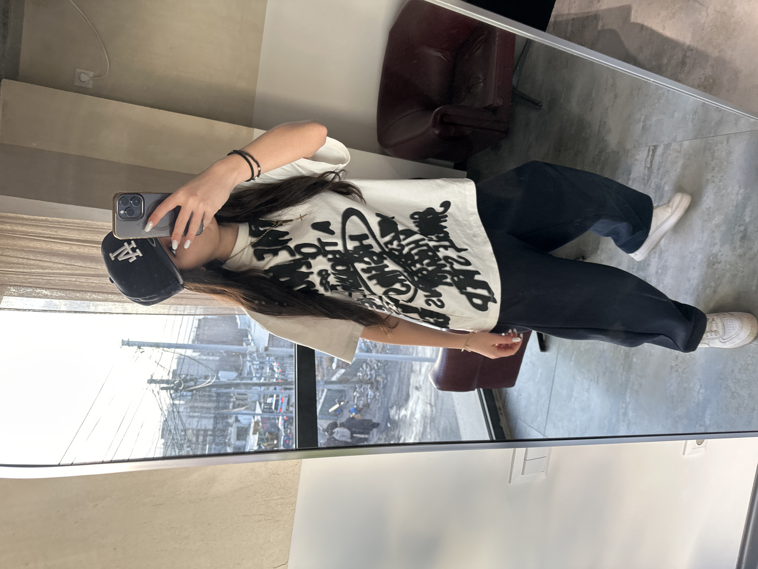 A woman wearing a black baseball cap, a white t-shirt with black graphics, and black pants takes a mirror selfie. She holds a phone in her hand. The mirror reflects a city street scene, a brown leather chair, and the woman's reflection. The floor appears to be tiled with a light gray finish. She also has on white sneakers.