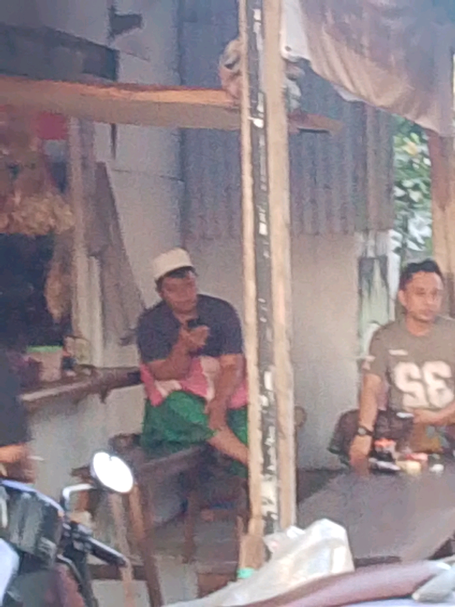 The image depicts an outdoor scene, possibly a market or roadside stall, in Probolinggo, Indonesia. Two men are visible. The man on the left is wearing a white cap, a dark shirt, and a patterned sarong. He is seated and holding a mobile phone. The man on the right wears a grey t-shirt with the number "35" on it and appears to be looking to his left. The background consists of corrugated metal walls, wooden structures, and some foliage. A motorcycle is partially visible in the foreground on the left. The overall impression is of a casual, everyday setting.