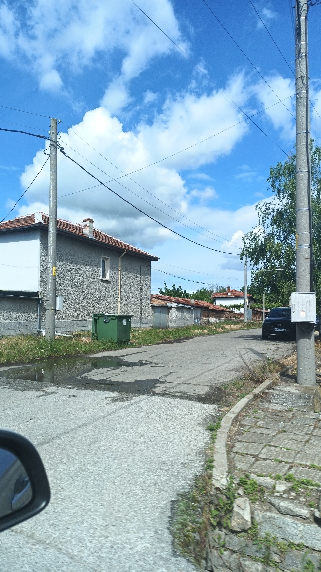 A street in Boliartsi, Bulgaria. On the left is a stone building with a red tile roof. Next to the building is a green dumpster. A puddle is on the road in front of the building. Further down the road is a black car parked next to a telephone pole. The sky is blue with clouds. Telephone wires are visible above the street. A car's side mirror is visible in the foreground.