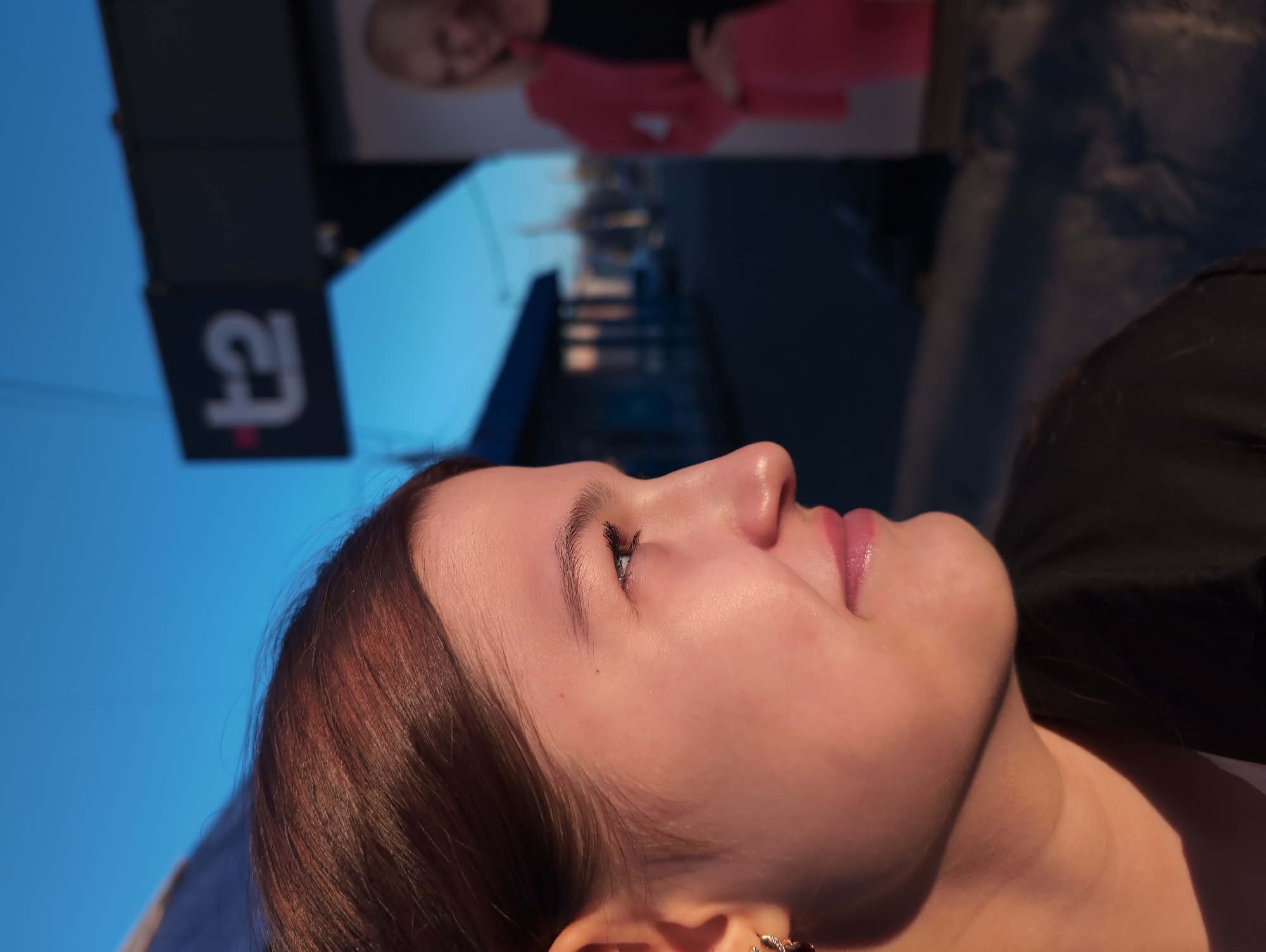 A close-up, angled view captures a young woman with fair skin and light brown hair gazing upwards. She has an earring in her left ear. The backdrop features a clear blue sky and a commercial sign with the number "2" and a logo, potentially a building or advertisement in the background. The location is possibly Izhevsk, Russia, based on contextual clues, but this cannot be definitively confirmed from the image itself.