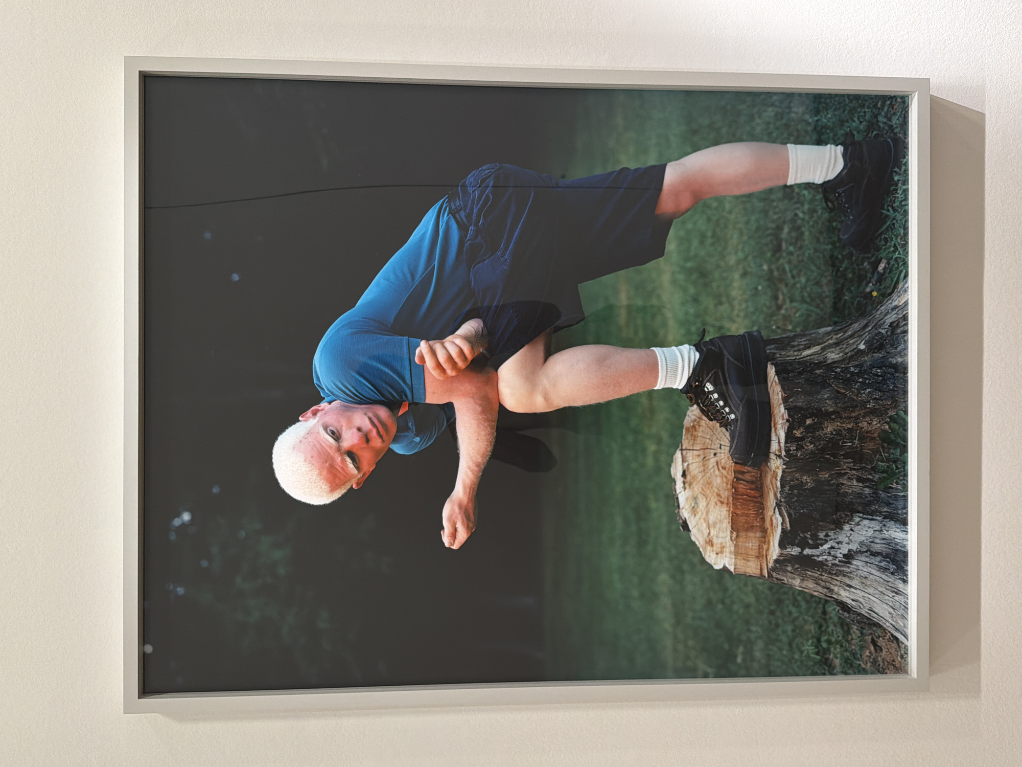 A framed photographic print, oriented horizontally, hangs on a light-colored wall. The photograph depicts a young person, possibly male, with remarkably white or platinum blonde hair, fair skin, and light eyes, consistent with albinism. He is wearing a short-sleeved blue t-shirt and dark blue shorts, along with white socks and black hiking-style shoes.
The person is captured in a dynamic, almost acrobatic pose within the photograph. One foot is firmly planted on the flat top of a weathered tree stump, while his other leg extends backward, creating a sense of balance or movement. His body is angled, and his arms are bent, with his hands positioned near his torso. He gazes directly forward towards the viewer with a neutral to slightly serious expression.
The background of the photograph is an outdoor setting, characterized by dark, muted greens, suggesting deep shade or a slightly underexposed forest or park area. A small patch of brighter green grass is visible towards the bottom right corner of the image, near the base of the tree stump. The lighting on the person is bright and clear, making him stand out against the darker background.
A notable detail is the vertical orientation of the person within the photograph, suggesting the photo might have been taken or displayed with a unique perspective, or perhaps the image is displayed sideways on the wall. The frame holding the photograph is a simple, light grey or silver color. There is no visible text within the photograph or on the frame. This media file is located in Paris, France.
