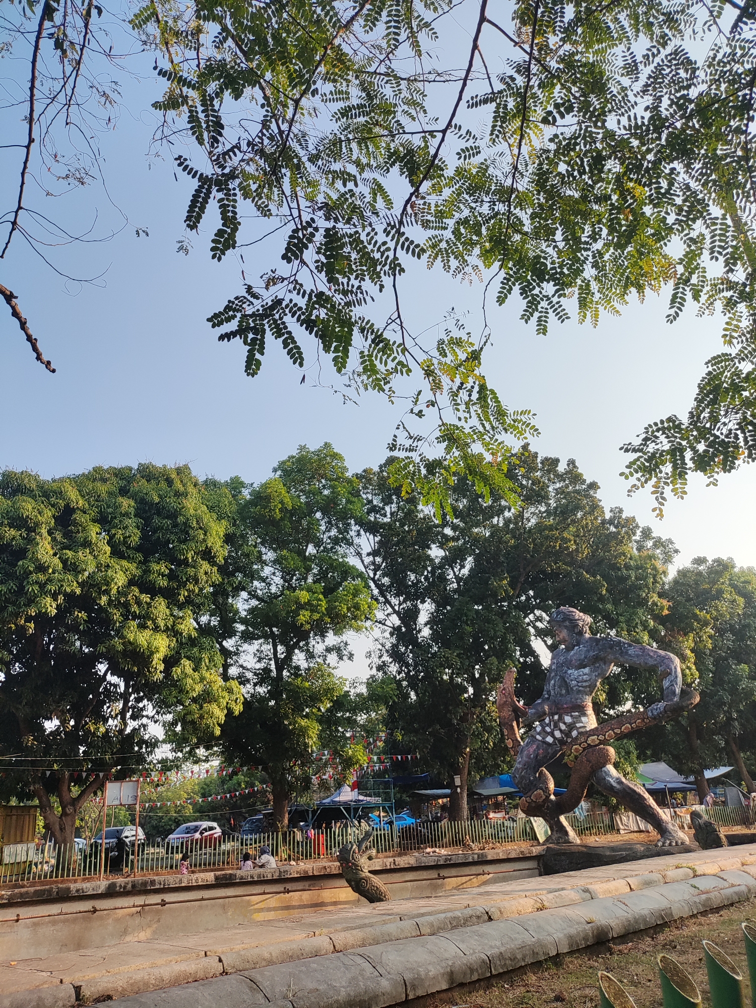 The image shows a statue of a man wrestling a snake in Cirebon, Indonesia. The statue is located in an outdoor area with trees and a clear sky. In the background, there are cars, tents, and people sitting near a fence. The statue itself is dark-colored and depicts a muscular male figure with a snake coiled around his body. The environment suggests a public park or recreational area.
