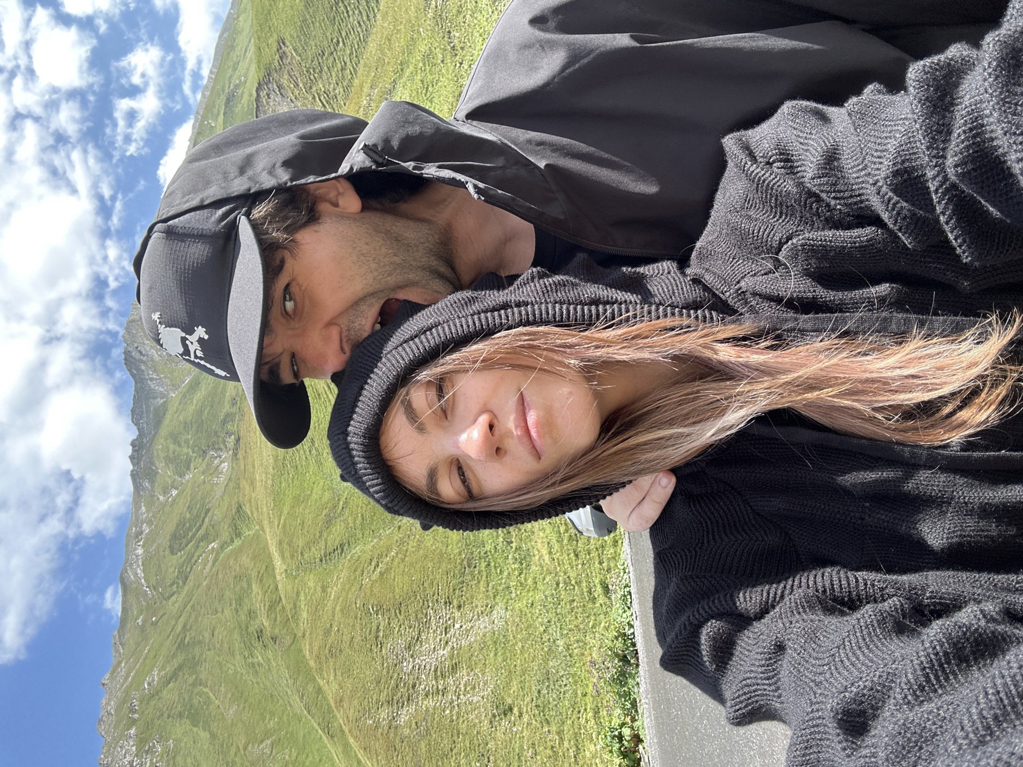 In this selfie, a couple is positioned in the foreground, with a lush green mountain range and a cloudy sky forming the background. The woman is on the left, facing forward with her head slightly tilted. She is wearing a black hooded sweater, and her hair is a light brown with hints of pink. Her expression is neutral, with her eyes open and looking at the camera. The man is on the right, leaning towards the woman and appears to be playfully nipping at her shoulder. He is wearing a black hooded jacket and a black baseball cap with a white emblem. His mouth is open in a wide grin, and he is looking at the camera. The mountain landscape behind them is characterized by steep, grassy slopes dotted with some rocky outcrops. The sky is partly cloudy with scattered white clouds against a blue backdrop. The setting appears to be a mountainous region, possibly along a road given the asphalt visible in the lower left corner. The specific location in Almasiani, Georgia, is not determinable from the visual information provided in the image.