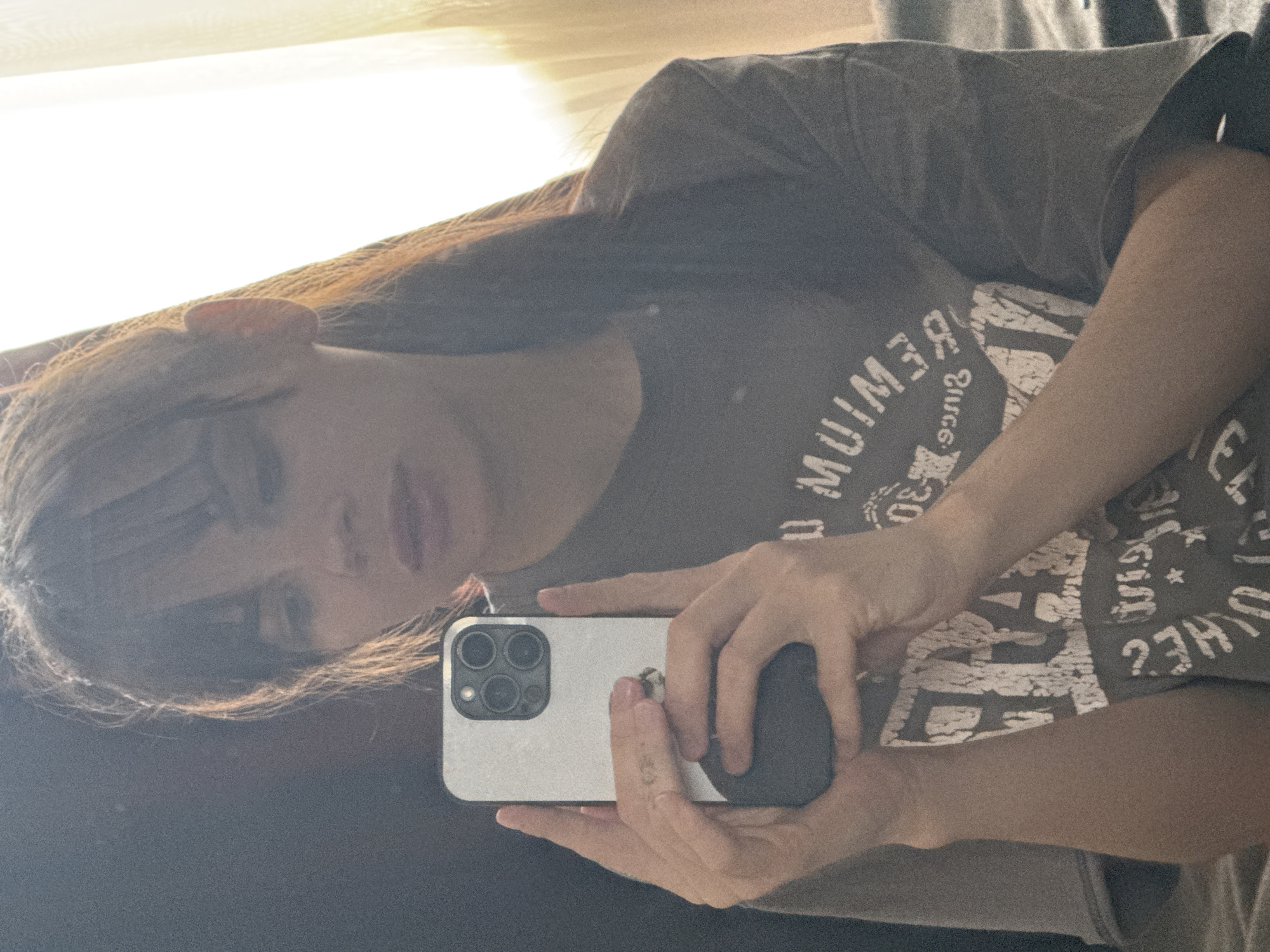 A young woman is taking a selfie with an iPhone. She is wearing a gray graphic t-shirt. Her hair is dark and partially covers her face. The background appears to be an interior with a blurred window. The location in the city of Rostov-on-Don, Russia, cannot be determined from the image.