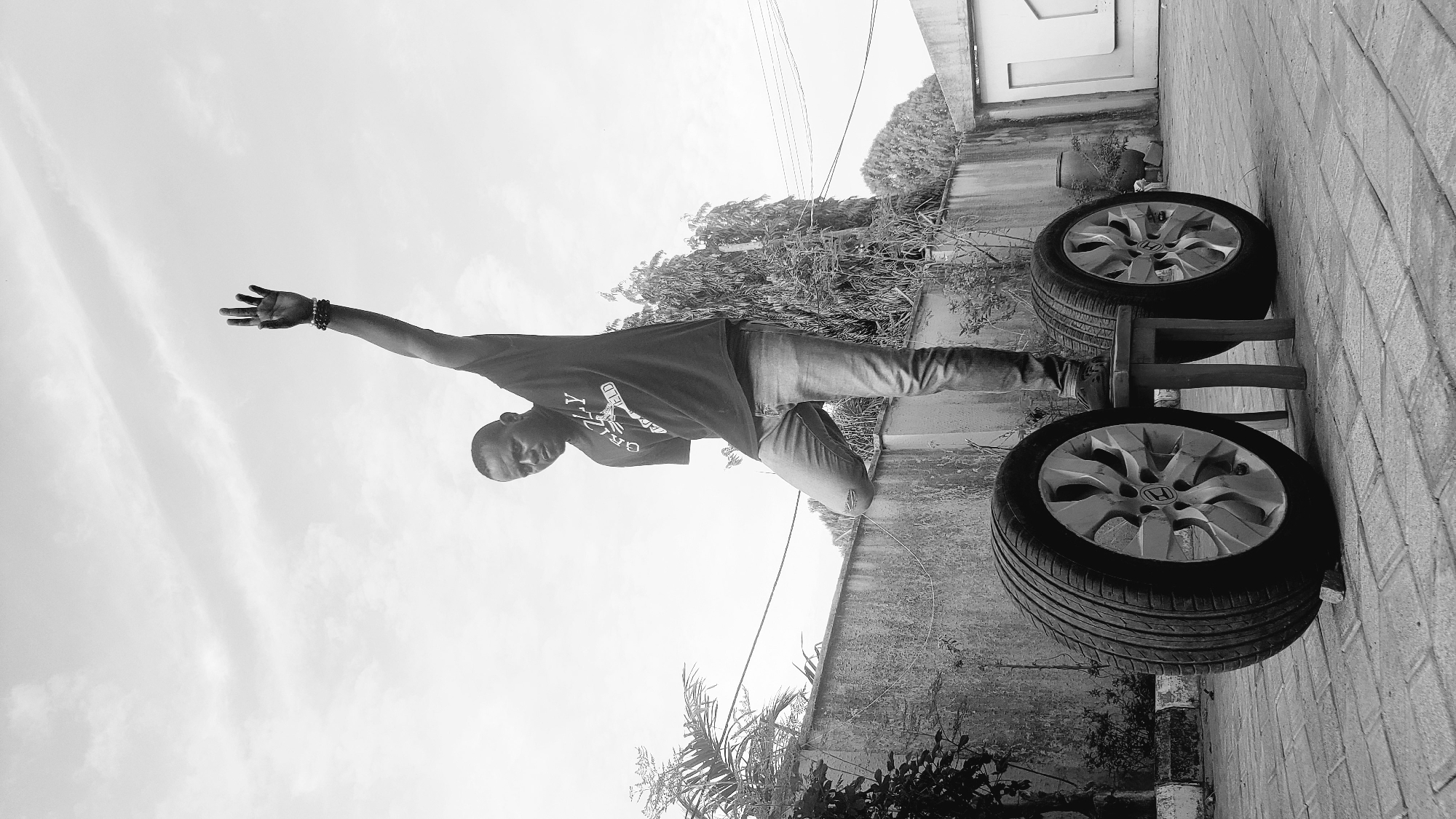 This black and white image, set in Sabon Tasha, Nigeria, depicts a young man posing outdoors during what appears to be bright daylight with an overcast sky. The man stands in the middle-left of the frame, facing slightly towards the camera with a neutral expression. He wears a dark t-shirt with the word "GRIZZLY" visible on the front, along with jeans that have a tear on the left knee. His left arm is extended upwards and outwards, with his fingers splayed, while his right arm is bent closer to his body. Multiple bracelets are visible on his left wrist.
To the right of the man, in the foreground, two car tires with distinct "H" (Honda) logo rims are stacked on a wooden stool or stand. They rest against a textured brick wall that forms the right boundary of the scene. Behind the man, a concrete wall stretches across the midground, with various bushes and sparse greenery interspersed along its base and extending upwards in the background. Overhead, a bright, cloudy sky dominates the upper portion of the image. Faint power lines are also visible crossing the background. The overall setting suggests a casual, outdoor area, possibly a backyard or a side alley of a building.