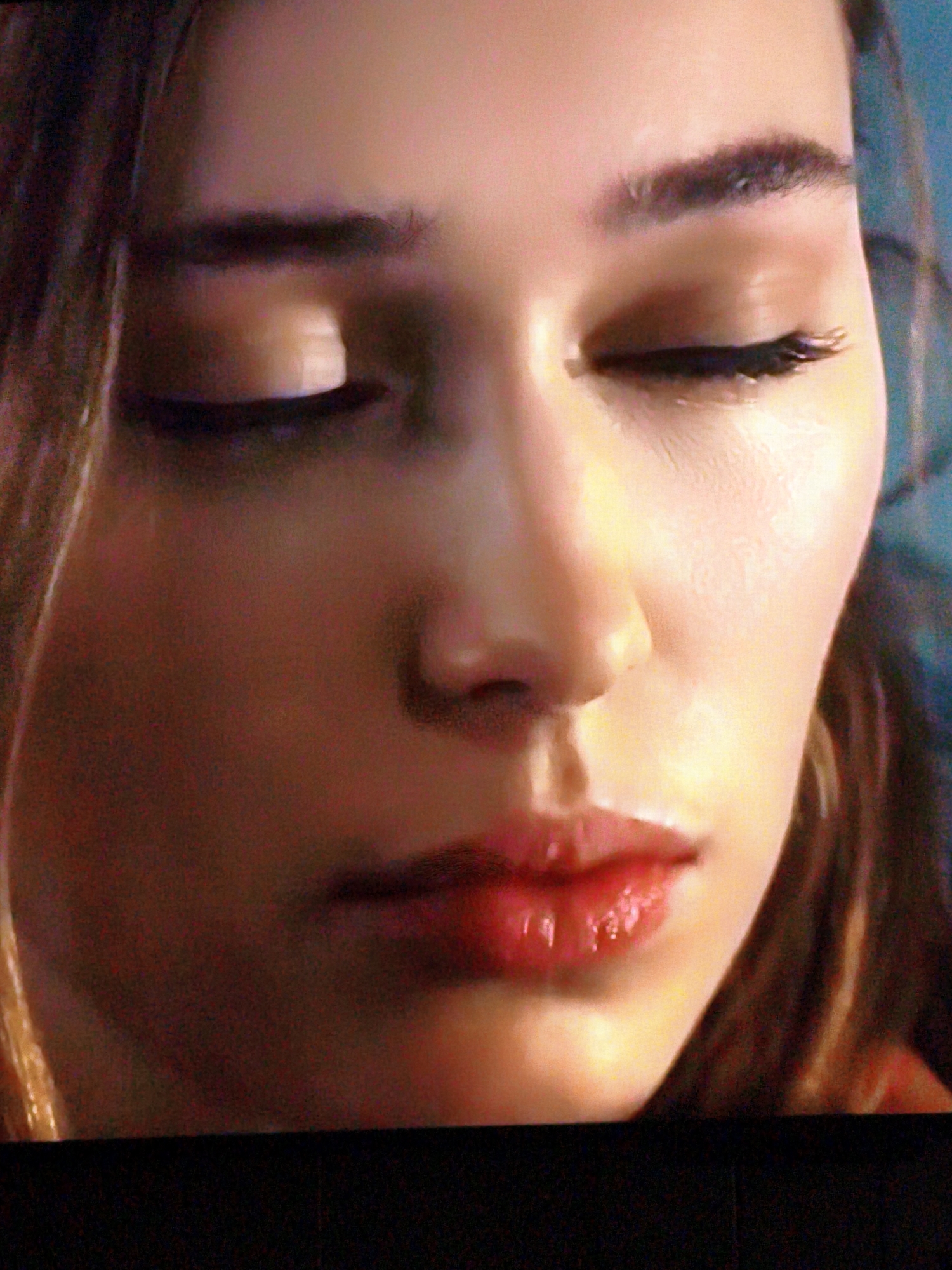 The image is a close-up, low-resolution photograph of a young woman's face with her eyes closed. Her lips are painted with a bright red lipstick that has a glossy finish. The lighting is warm, highlighting the contours of her face and the sheen on her lips. Her eyebrows are dark and well-defined. Her hair, a light brown color, frames the left side of her face. The background is blurred and appears to be a dark, possibly blue, color. The photograph is cropped tightly, focusing solely on the woman's facial features. There is no indication of any specific location in Vladivostok, Russia, within the image content.