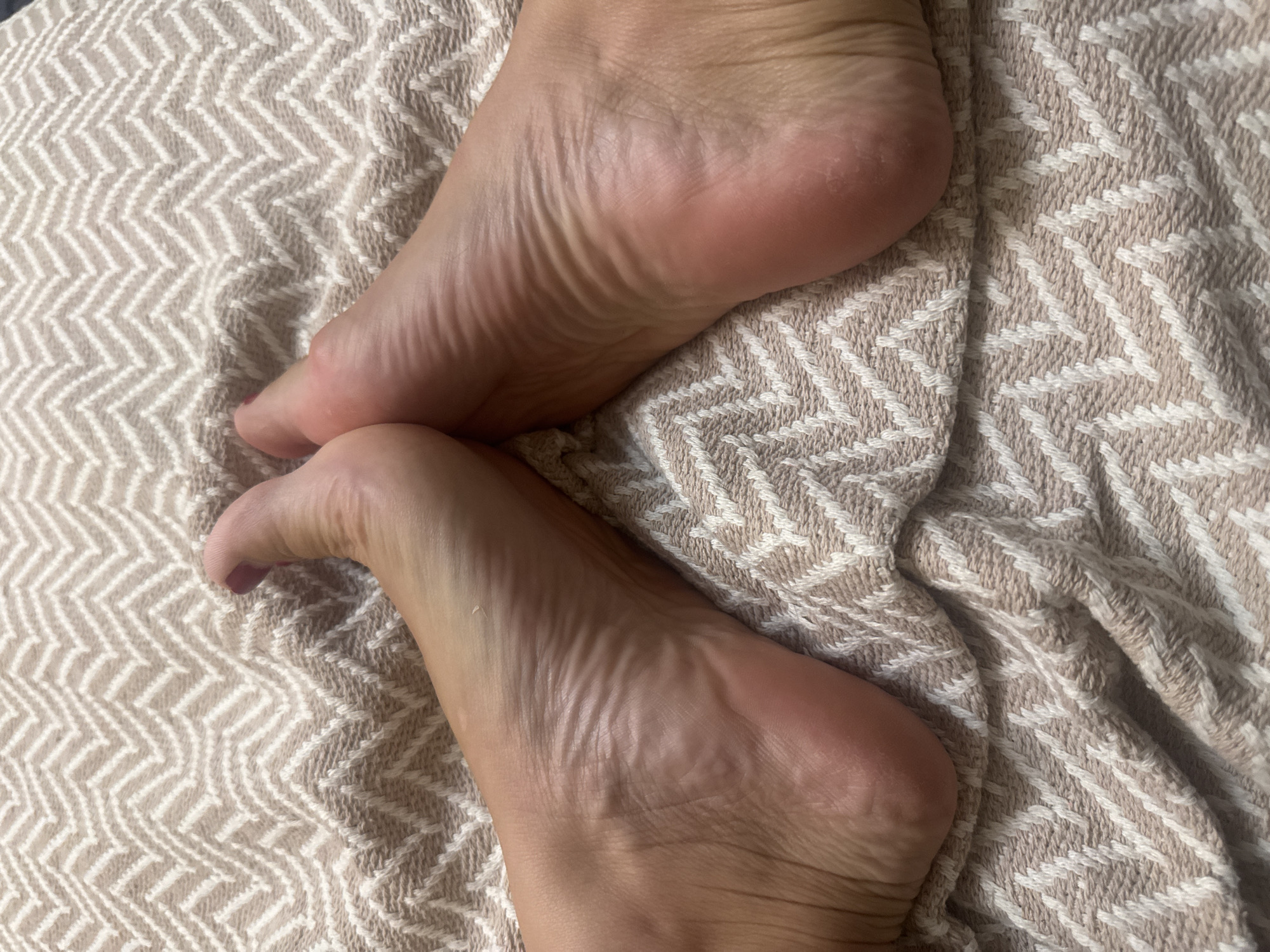 Two bare feet, facing each other with the soles visible, rest on a light-colored, chevron-patterned fabric. The feet are positioned in a way that the arches are prominently displayed. The fabric appears to be a blanket or throw. The scene is lit, and the image is a close-up, focusing solely on the feet and the fabric. There are no discernible details about the specific location within the city of Omsk, Russia, nor any actions or interactions taking place. The image is a static, neutral depiction of feet on fabric.