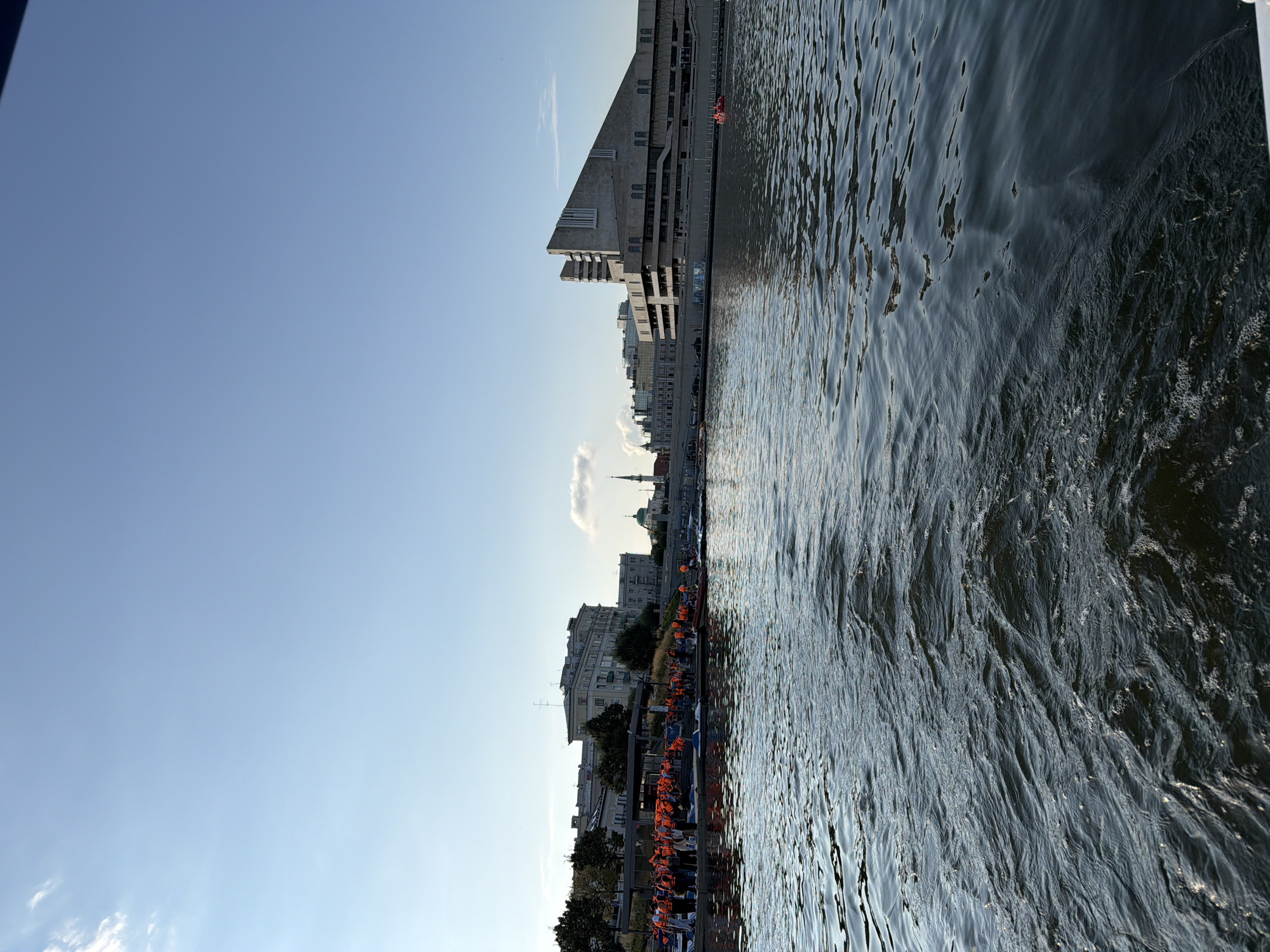 In Kazan, Russia, a clear sky with a few wispy clouds hovers over a body of water. In the foreground, the rippling water creates a dynamic texture. Along the embankment, a crowd of people wearing bright orange life vests is gathered, some appearing to be preparing for an activity. Various boats are moored at the dock, including what appear to be paddle boats. In the background, the cityscape unfolds with a mix of modern and older architectural styles. Notable buildings include a large, angular structure on the right and several multi-story buildings with distinct facades further left. A minaret and a green-domed building suggest the presence of religious structures within the urban landscape. The overall scene depicts a sunny day by the water, with a group of people engaged in an outdoor activity.