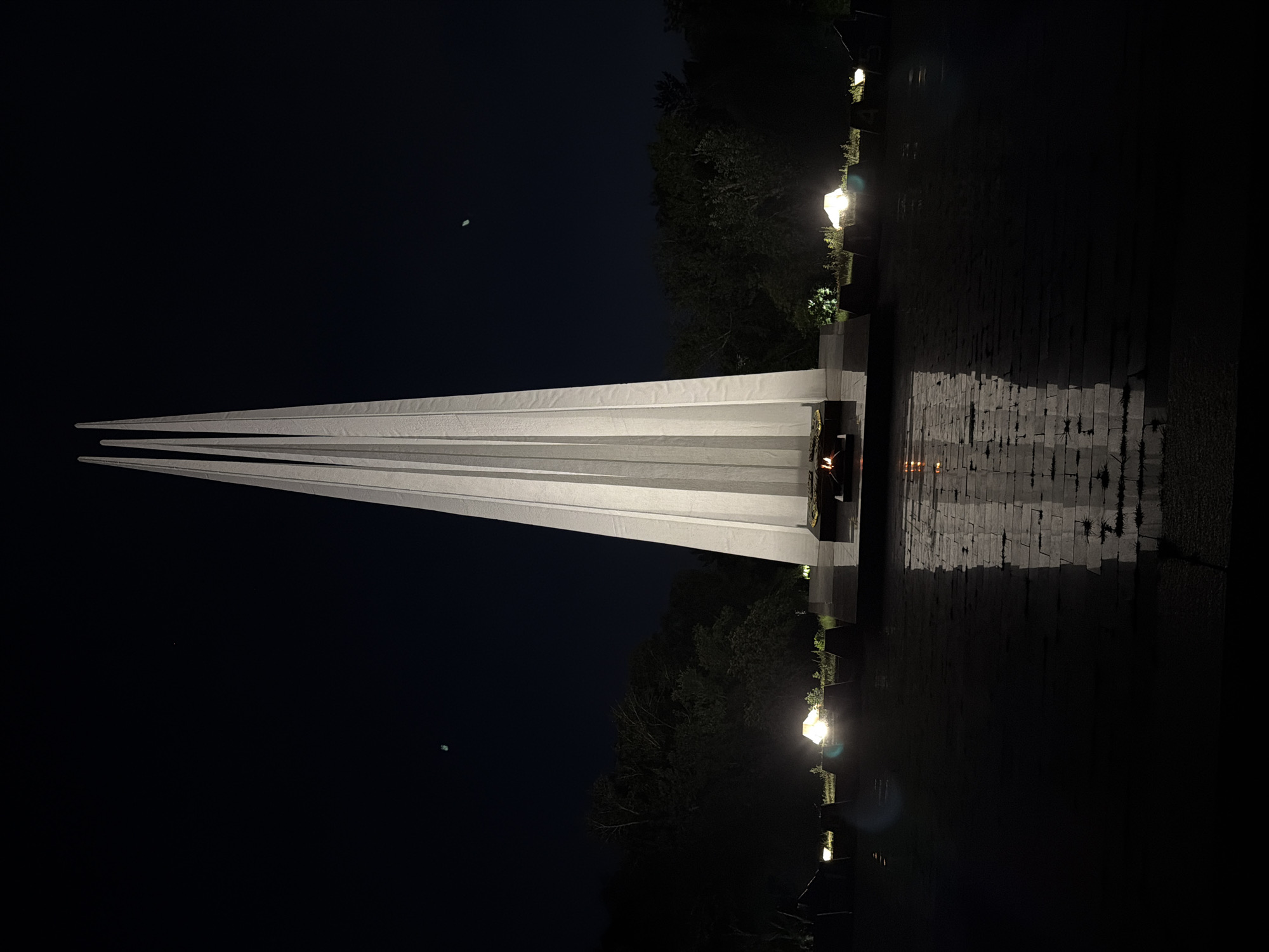 The image shows the Eternal Flame memorial in Pavlodar, Kazakhstan, at night. The memorial is a tall, white, spire-like structure with three pointed sections, illuminated from below. It stands on a dark, reflective surface, possibly water or polished stone, which mirrors the structure and the flames. In front of the spire, an eternal flame burns. The background consists of dark trees and a deep blue night sky. Several small, bright lights are visible around the base of the memorial, illuminating the surrounding area. There are also two small, faint celestial bodies visible in the sky. The overall scene conveys a solemn and commemorative atmosphere.