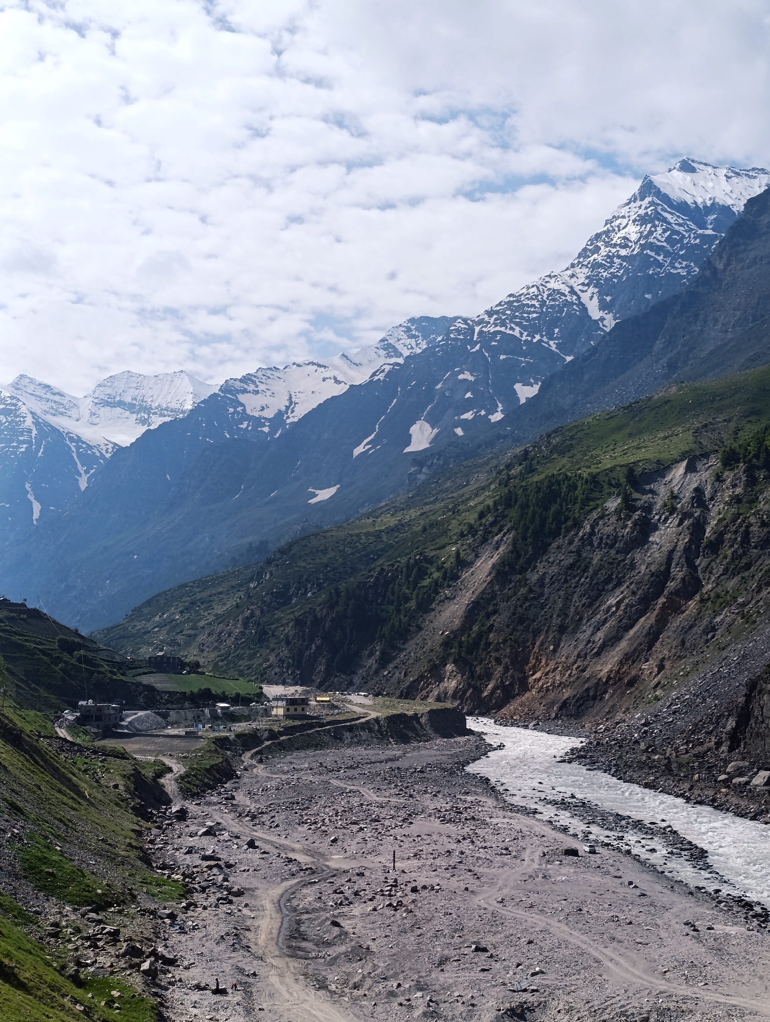 A vast, rugged mountain landscape dominates the image, with a wide, rocky riverbed in the foreground. Snow-capped peaks pierce the sky in the distance, their stark white contrasting with the deep blue of the mountains and the lush green of the lower slopes. A turbulent river, its water a milky white, snakes its way through the valley, its banks lined with loose stones and gravel.
In the mid-ground, a small cluster of buildings, likely part of the town of Sissu in India, is nestled against the mountainside. A dirt road winds its way through the rocky terrain, providing access to the settlement. Several vehicles are visible, including what appear to be SUVs, suggesting some level of activity and transportation within the remote area.
The sky above is a canvas of white, puffy clouds scattered across a pale blue expanse, hinting at a bright, possibly cool day. The overall impression is one of dramatic natural beauty, with human presence appearing minimal and subservient to the imposing power of the mountains and the river.