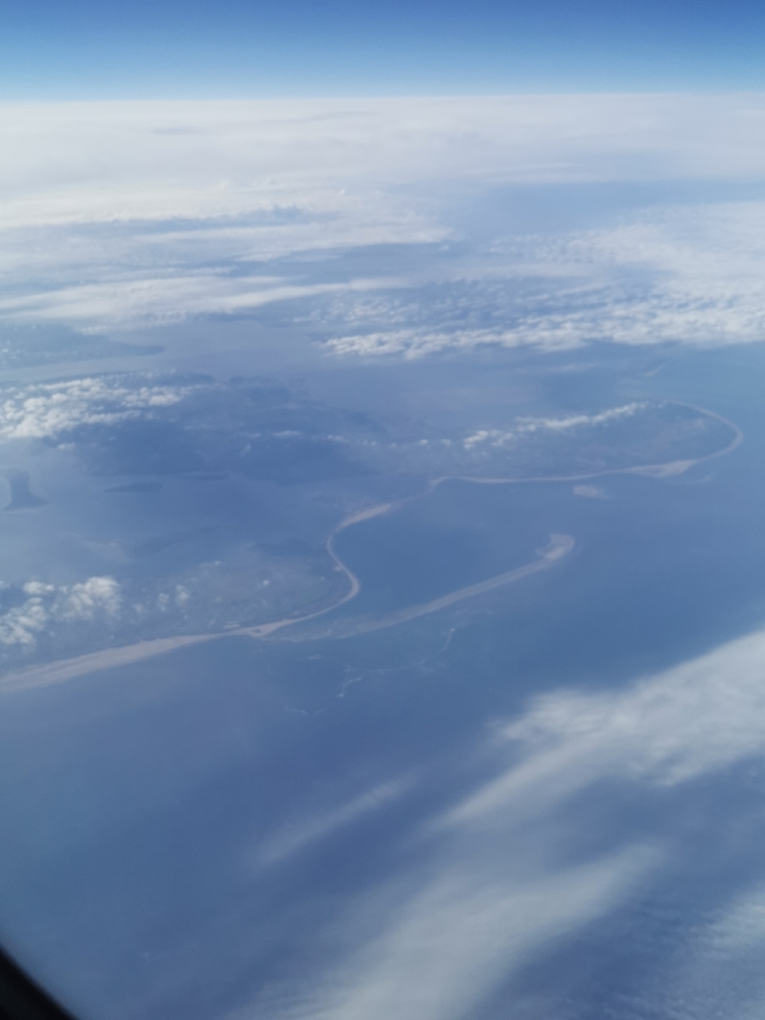 An aerial view from an airplane shows a landscape of water and land formations under a cloudy sky. The scene presents a blue sea intersected by thin strips of land. The lighting suggests daylight conditions. The composition of clouds above suggests altitude and perspective from the plane.
Given the land formations are in Varna, Bulgaria, the picture might be of the Black Sea near the Bulgarian coast.