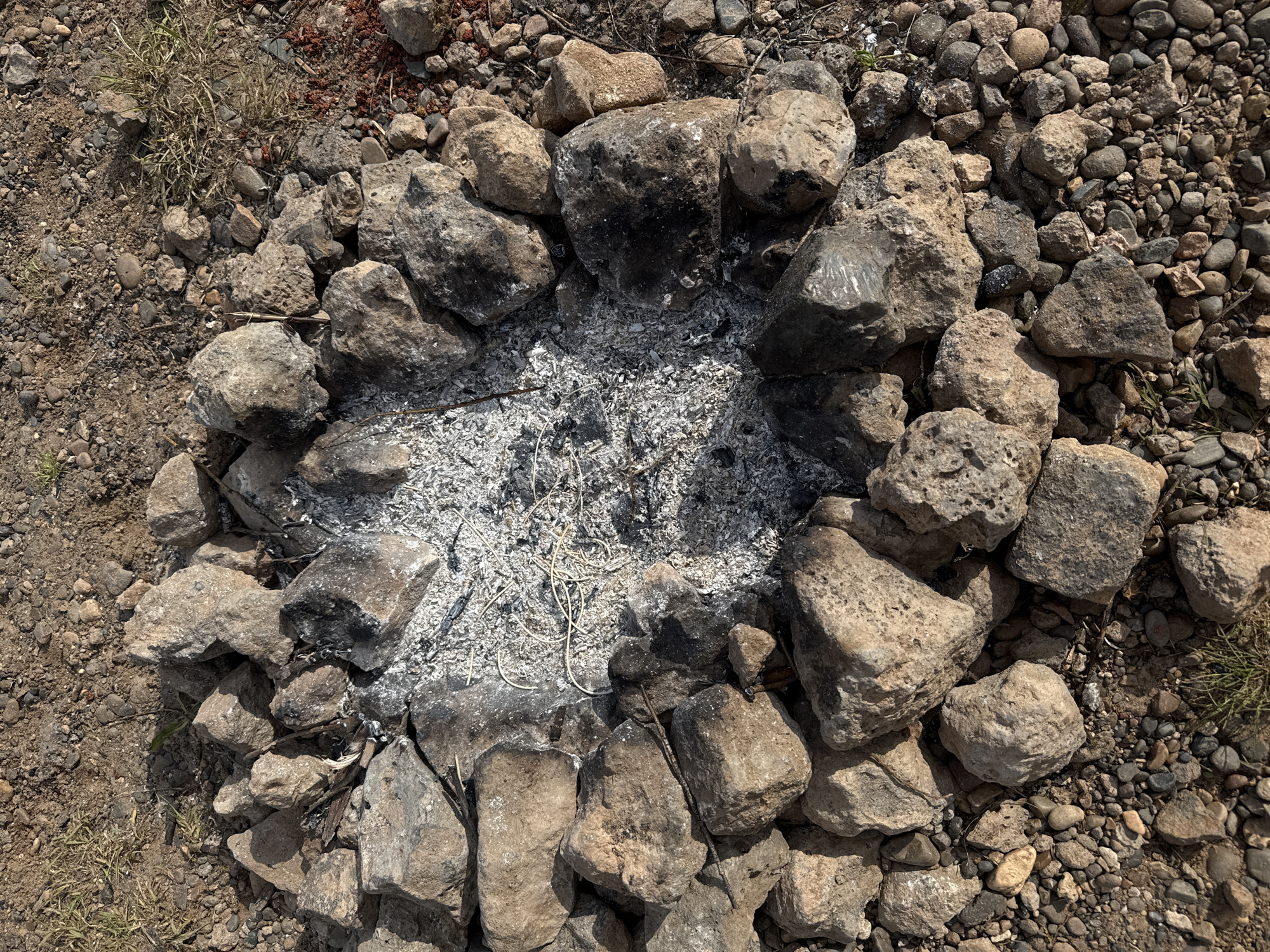 The image shows a circular fire pit constructed from roughly hewn rocks and stones. The interior of the pit contains ash and charred remnants of burned material. The surrounding ground is a mixture of soil, gravel, small stones, and sparse vegetation. The location is unspecified, and there is no indication of human presence in the image. The scene depicts a fire pit in a natural or semi-natural environment. There is no evidence to indicate the location as Povarenki, Russia.