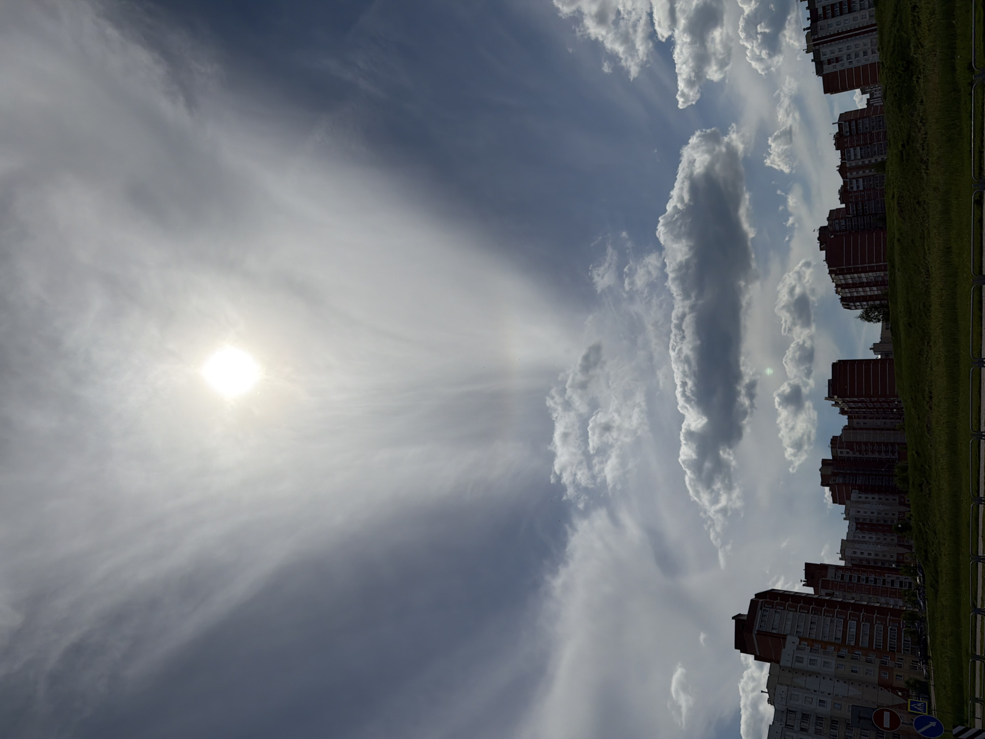 The image captures a cityscape under a sky dominated by scattered clouds, sunlight, and a hazy atmosphere. Tall apartment buildings line the horizon, their facades featuring rows of windows. A green strip of land is visible. The sun shines brightly in the sky. The picture is taken from a tilted perspective.