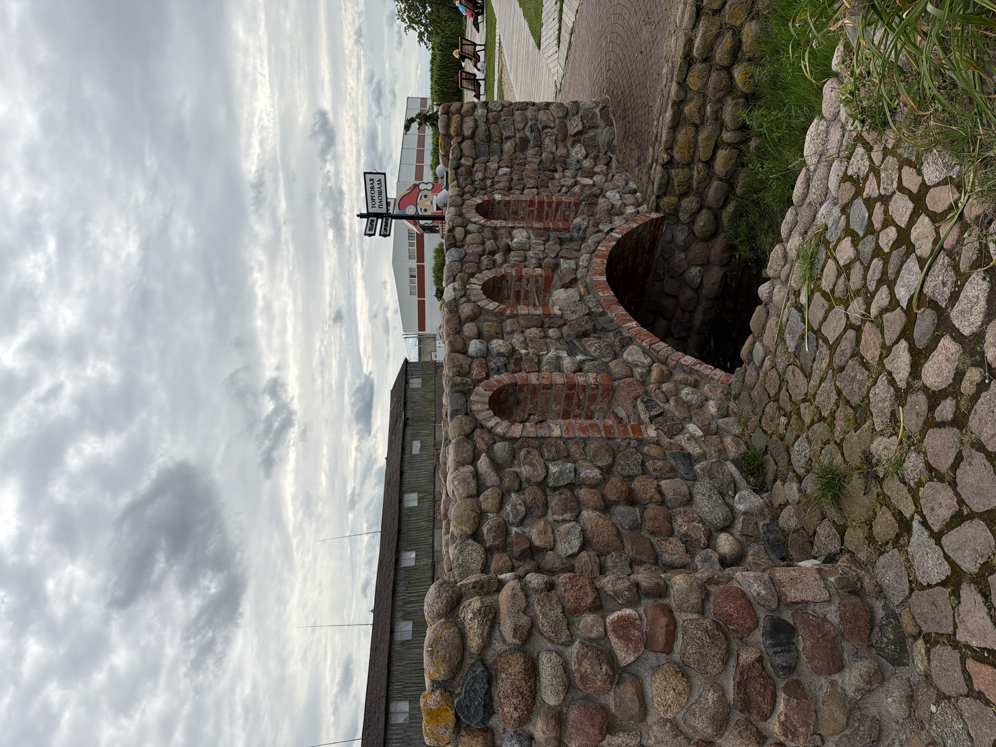 This image depicts a small, rustic stone bridge in Nekrasovo, Russia. The bridge is constructed from rough, irregularly shaped stones of various colors, with arched openings. The area around the bridge is paved with cobblestones, and there is vegetation growing in the crevices and along the sides. In the background, a long wooden building with windows is visible, along with a modern white building with a red stripe. A directional signpost points towards "ТОРГОВАЯ ПЛОЩАДЬ" (Trade Square) and "ПЕКАРНЯ" (Bakery). A colorful, stylized statue of a person's head is positioned behind the signpost. Further in the background, people are seated on benches. The sky is overcast with heavy clouds.
