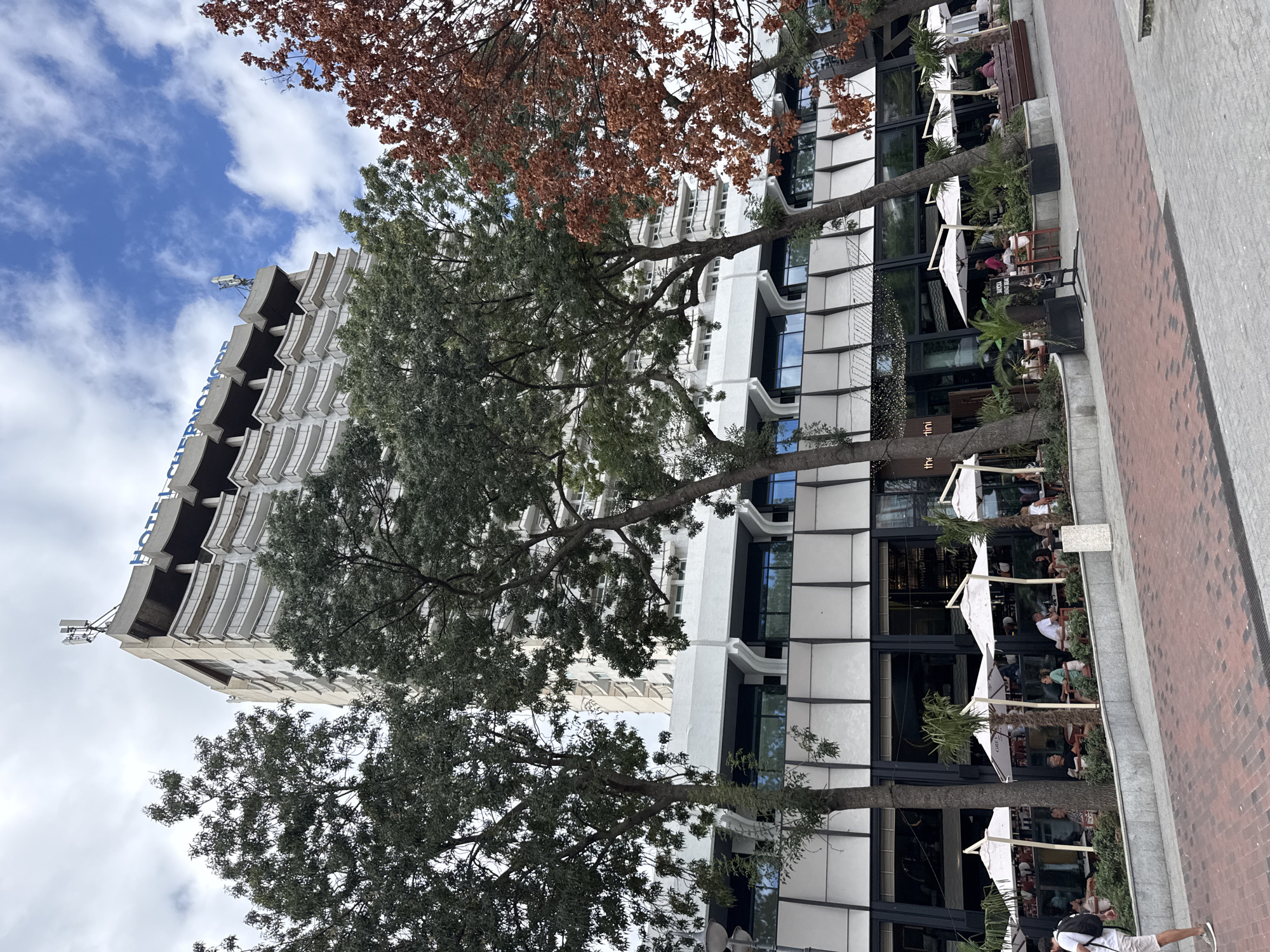 The image shows a wide view of the Hotel Chernomore in Varna, Bulgaria, on a cloudy day. The hotel is a tall, white, multi-story building with many balconies. In front of the hotel is a paved area with tables and umbrellas set up for outdoor dining at a restaurant. Several people are seated at the tables, some eating and others talking. Trees with green and reddish-brown leaves are scattered throughout the scene, some leaning over the outdoor seating area. The ground is paved with red and gray bricks. A person in a white t-shirt and light-colored shorts is walking away from the camera on the left side of the image.