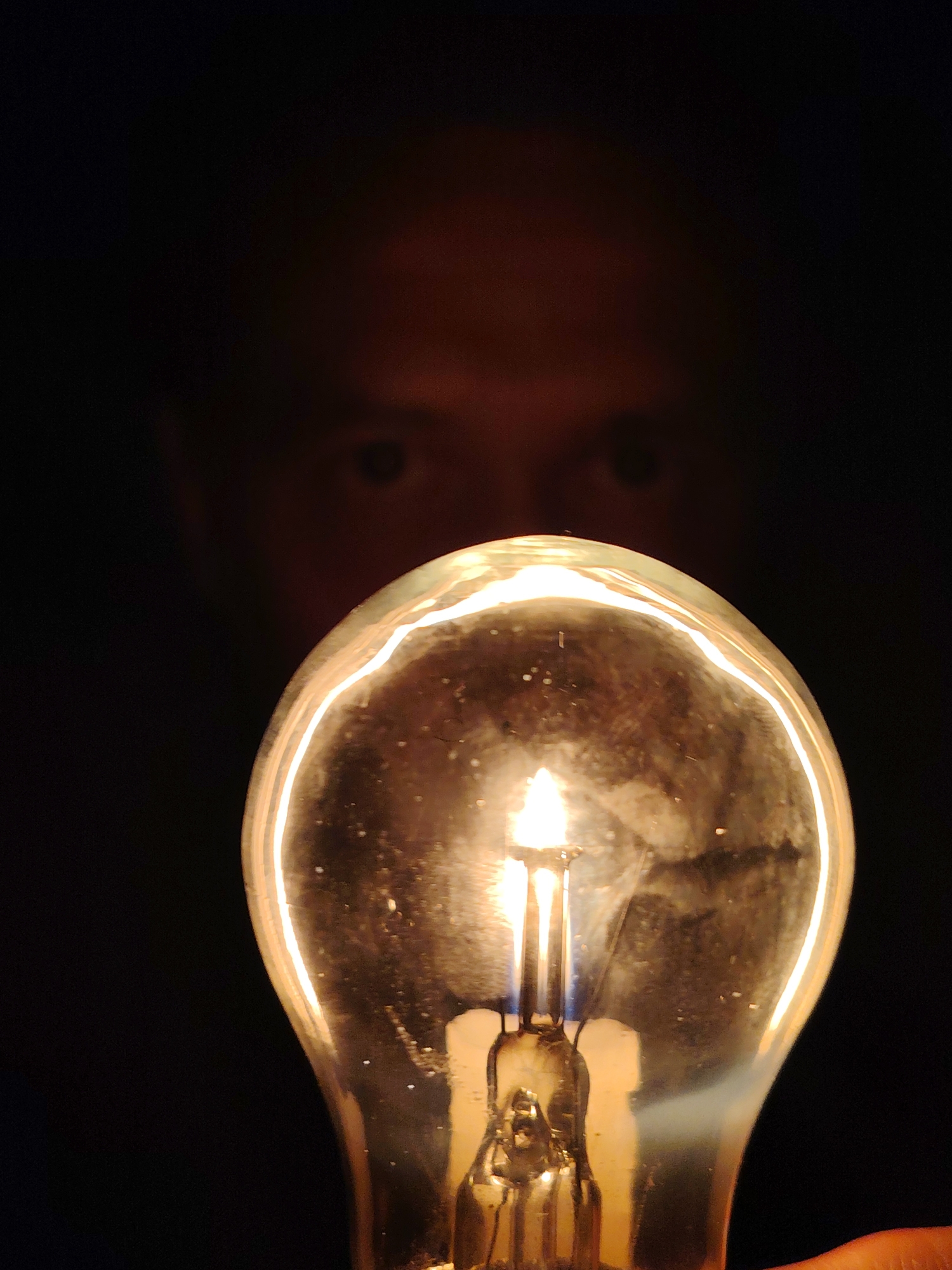 A close-up, low-angle shot focuses on a lit incandescent light bulb against a dark background. The bulb, the primary subject, is positioned in the foreground and appears to be held or supported by a hand, partially visible at the bottom right. The filament within the bulb is clearly illuminated, casting a warm glow. Behind the bulb, the blurred silhouette of a person's face is discernible, with two eyes visible in the shadows. The overall lighting is dim, with the bulb serving as the main light source, creating a dramatic contrast between the illuminated foreground and the dark background. The image does not contain any identifiable landmarks or elements that would indicate its location in Chaikovsky, Russia.