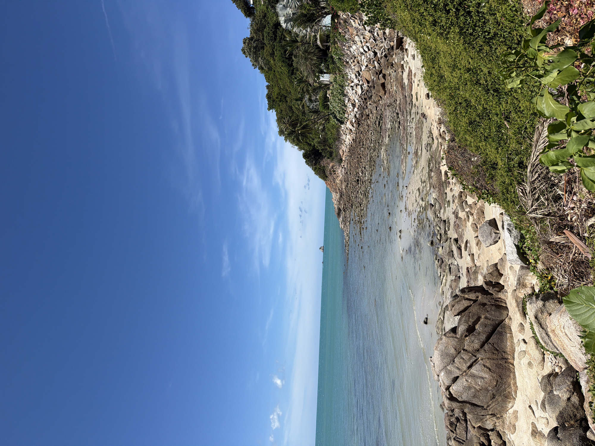 The image depicts a rocky coastline in Baan Plai Laem, Thailand, under a clear blue sky with scattered wispy clouds. The sea, a vibrant turquoise, gently laps against a shore littered with numerous rocks and pebbles of varying sizes. In the distance, a small island or rock formation emerges from the water, and a small boat is visible on the horizon.
The land slopes upwards from the shore, covered in lush green vegetation, including palm trees and dense bushes. Several small white structures, possibly houses or resort buildings, are nestled within the greenery on the hillside. Large boulders are present on the sandy and rocky beach in the foreground. The overall scene is one of a serene tropical coastal environment. There are no people visible in the image.