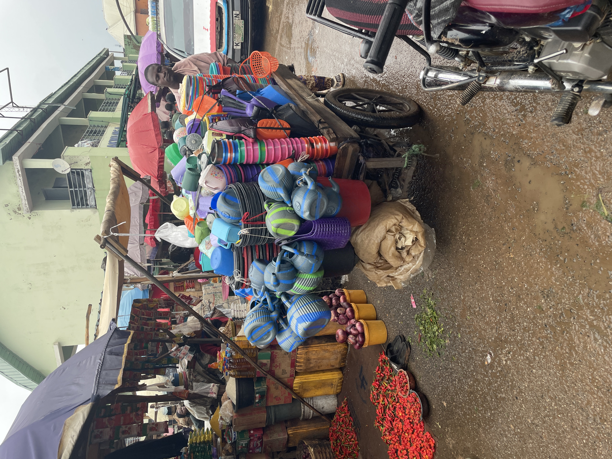The image depicts a busy market scene in Akwanga, Nigeria, likely during the daytime. The ground is wet and muddy, suggesting recent or ongoing rain, contributing to a damp and overcast atmosphere. A man, presumably a vendor, is visible in the upper left portion of the image, wearing a light-colored shirt. He is surrounded by a large assortment of colorful plastic wares, including buckets, basins, and containers, stacked on a cart or stall. Red and purple umbrellas are also present, adding to the market's vibrant yet slightly chaotic ambiance.
Further into the scene, various goods are displayed, including red chilies, onions in yellow containers, and other food items. The presence of a motorcycle further indicates the bustling nature of the market and the mode of transportation. The background shows a multi-story building, suggesting an urban or semi-urban market setting. The overall impression is one of a lively marketplace with a wide variety of goods being offered for sale, set against the backdrop of a wet, possibly humid, environment typical of a rainy day in Nigeria. No distinct text is clearly legible in the image.