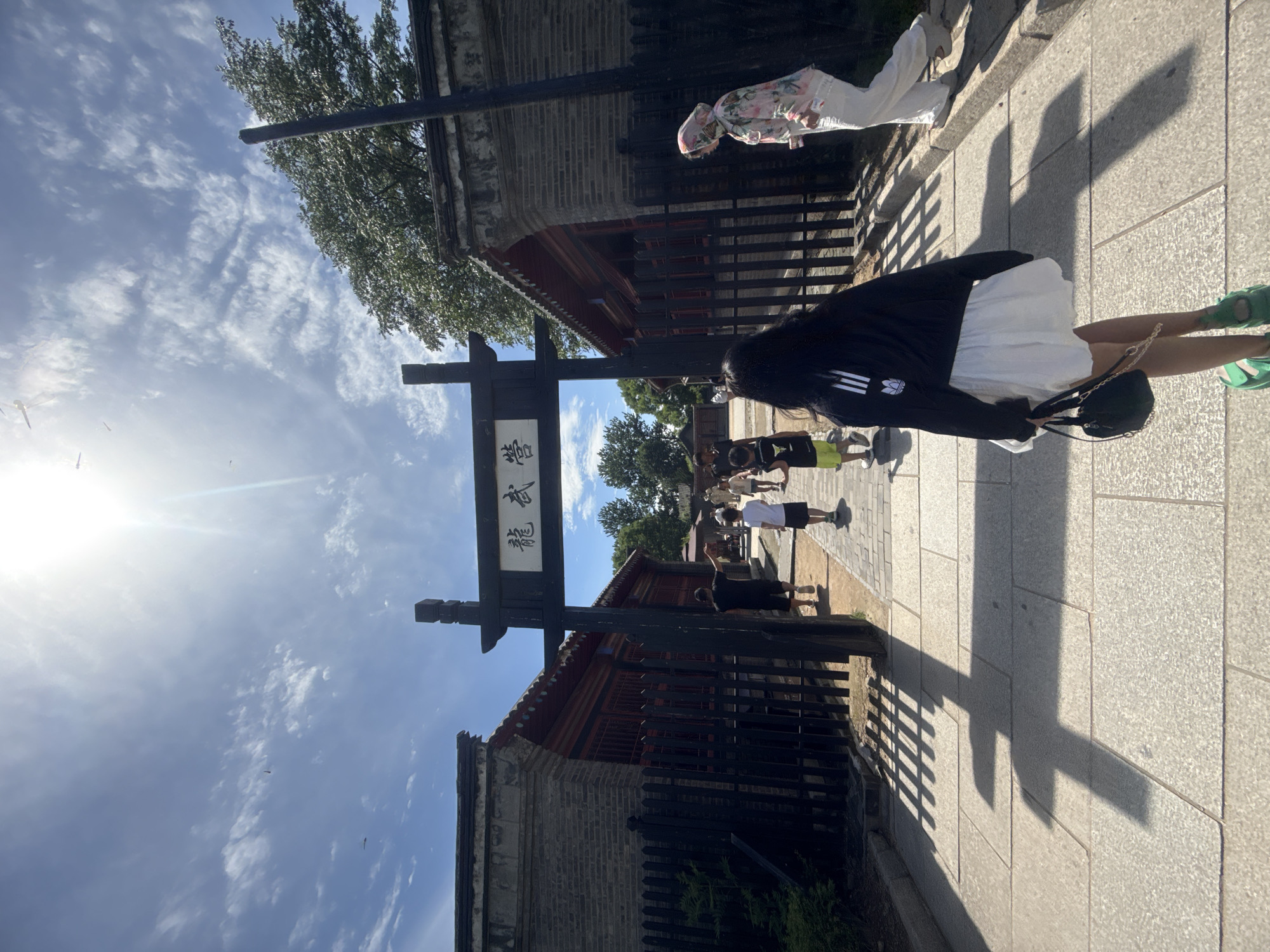 In Qinhuangdao, China, a group of people are walking towards an ornate wooden gate with Chinese characters, possibly signifying a temple or historical site. The sun shines brightly from behind scattered clouds, casting long shadows on the stone pathway. The architecture suggests traditional Chinese design, with dark wood and grey brick buildings on either side of the entrance. A young woman in a black hoodie and white skirt is in the foreground, facing away from the camera, holding a black handbag. Other individuals, including children, are walking ahead of her. A woman in a floral hijab and white pants is seen on the right side of the frame, walking away from the viewer. The overall atmosphere is one of a sunny day at a cultural or historical location.
