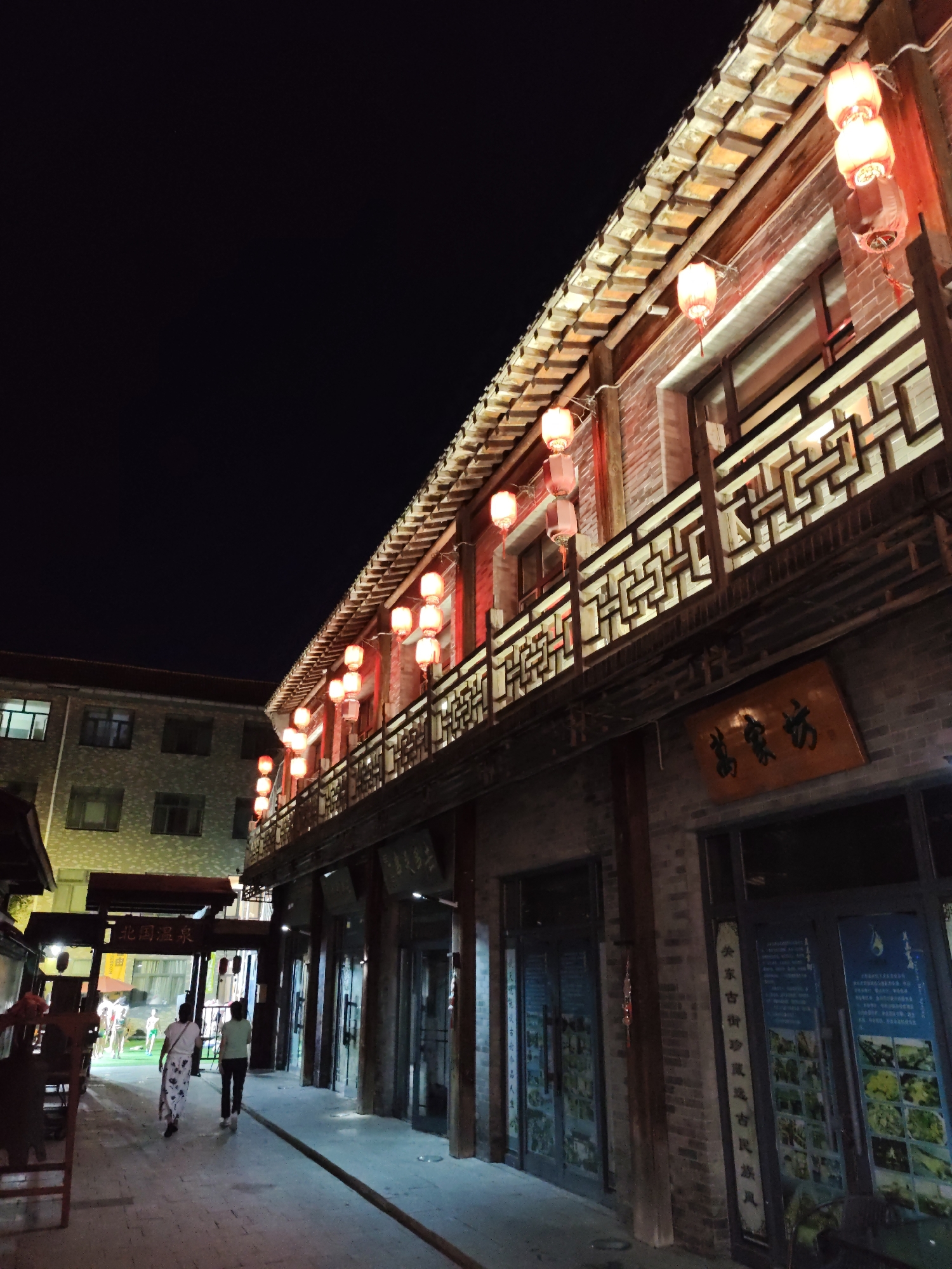 In Lindian, China, at night, a street scene unfolds with traditional architecture illuminated by red lanterns. The main building, constructed of brick with wooden accents, features a multi-tiered roof and an ornate railing on the upper level, from which numerous red lanterns hang. Below the upper level, a wooden sign with Chinese characters is visible. Adjacent to this building, a modern structure with multiple windows is seen. Two individuals walk away from the camera down a paved walkway lined with shops. One person wears a white top and a patterned skirt, while the other is dressed in a green top and dark pants. A red sign with Chinese characters hangs above an archway. The overall atmosphere is one of a lively commercial street at night.