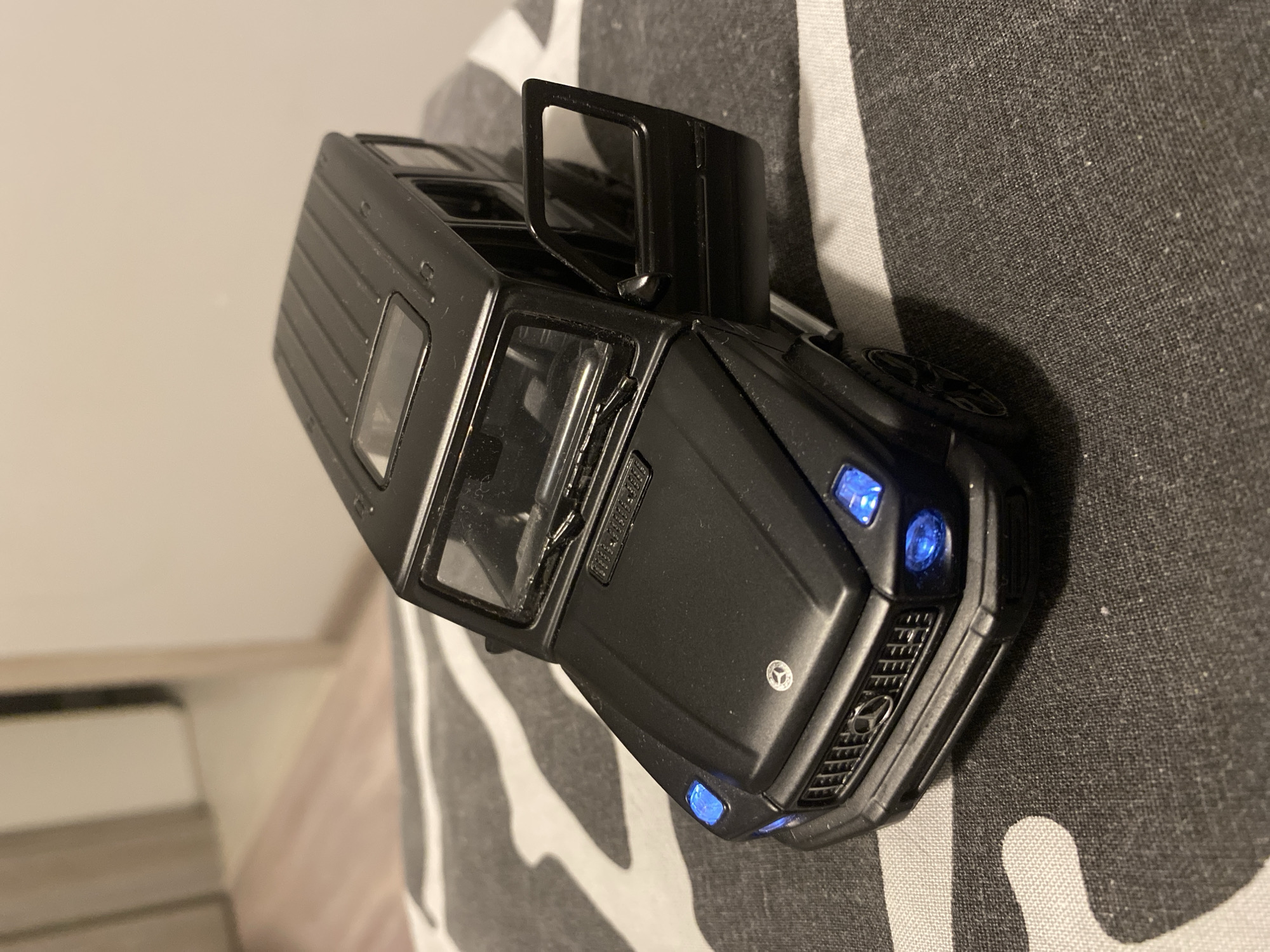A black toy Mercedes-Benz G-Wagon is positioned on a striped, textured surface. The toy car is detailed, with working doors and illuminated blue headlights. The background appears to be an indoor setting, possibly a room with wooden flooring and a doorway visible, suggesting a domestic environment. There is no indication of the city of Łódź, Poland.