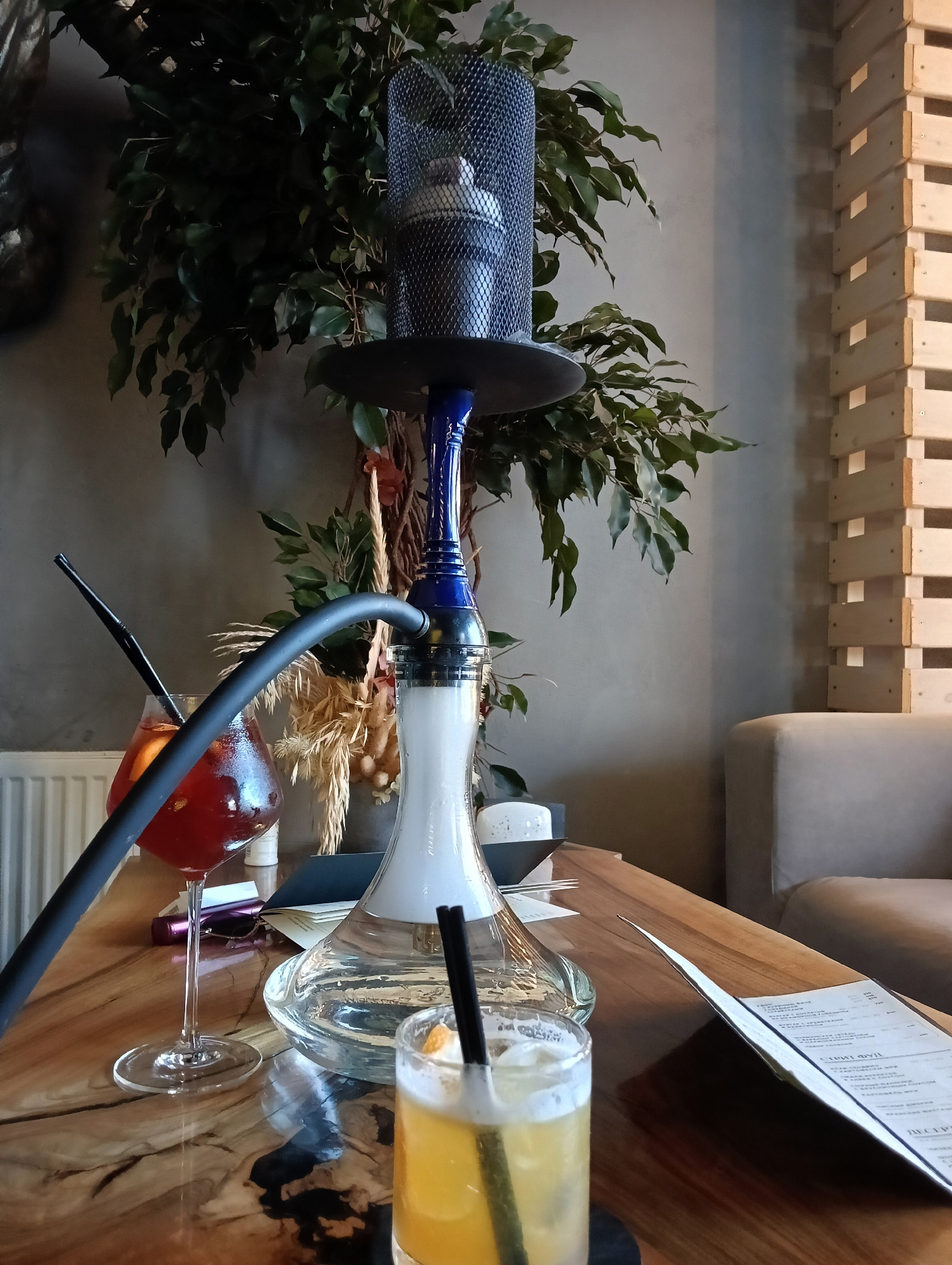 The image depicts a dimly lit interior of a hookah lounge or bar, likely located in Saint Petersburg, Russia, given the context of the request. The primary focus is a modern hookah setup, predominantly blue and black, placed on a wooden table. Adjacent to the hookah, on the left, is a stemmed glass filled with a reddish-orange beverage and a black straw. Another shorter glass with a yellow beverage and a straw is positioned in the foreground. A menu lies open on the table to the right, with text in Cyrillic script. The background features a large potted plant with dark green leaves, a textured grey wall, and a wooden decorative structure made of stacked planks on the right. A light-colored sofa is partially visible in the far right background. There are no people visible in the image. The lighting is ambient and somewhat muted, creating a relaxed atmosphere.