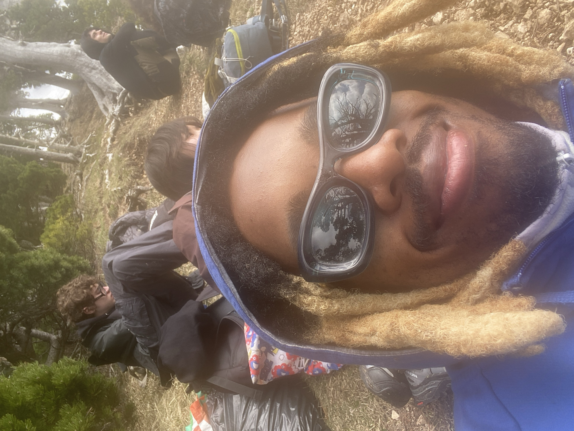 This sideways-oriented selfie captures a group of people resting outdoors, likely during a hike or outdoor excursion in a natural setting. The location context of Chichilianne, France, suggests a mountainous or rugged landscape.
The primary subject, prominently featured in the foreground, is a person with dark skin, distinctive light brown/blonde dreadlocks, a mustache, and a beard. They are wearing a blue hooded jacket and dark, reflective sunglasses. The sunglasses reflect a bright, clear sky with clouds and the intricate, bare branches of trees, indicating daylight and good weather. The person has full lips and a relaxed expression, with a hint of a smile, looking directly at the camera. Their left foot, adorned with a dark hiking shoe bearing the visible brand name "SALOMON," is seen at the bottom right.
In the background, several other individuals are visible. To the upper left, a person in a dark hooded top is seated or crouching, looking upwards. Further to the lower left, another person with curly brown hair and glasses is also resting. Backpacks, including a grey and green one, are scattered around, along with black plastic bags and a colorful patterned cloth on the ground, indicating outdoor activity gear.
The surrounding environment features dry grass, dirt, and rocks underfoot, with a scattering of trees, many appearing as bare trunks and branches, suggesting an environment typical of higher altitudes or perhaps an early spring or late autumn season. Some green foliage is visible on the far left. The overall scene conveys a moment of rest and enjoyment in a rugged outdoor environment during the daytime.